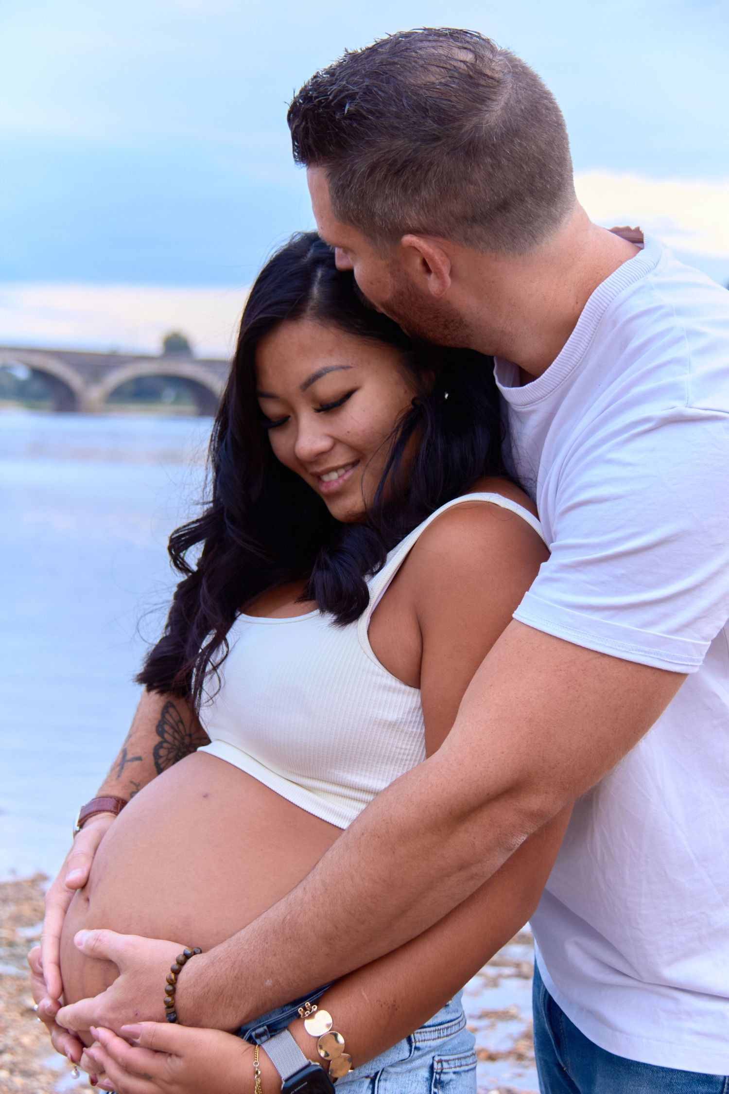 A tender maternity photo showing an expectant couple embracing by a waterfront at sunset.