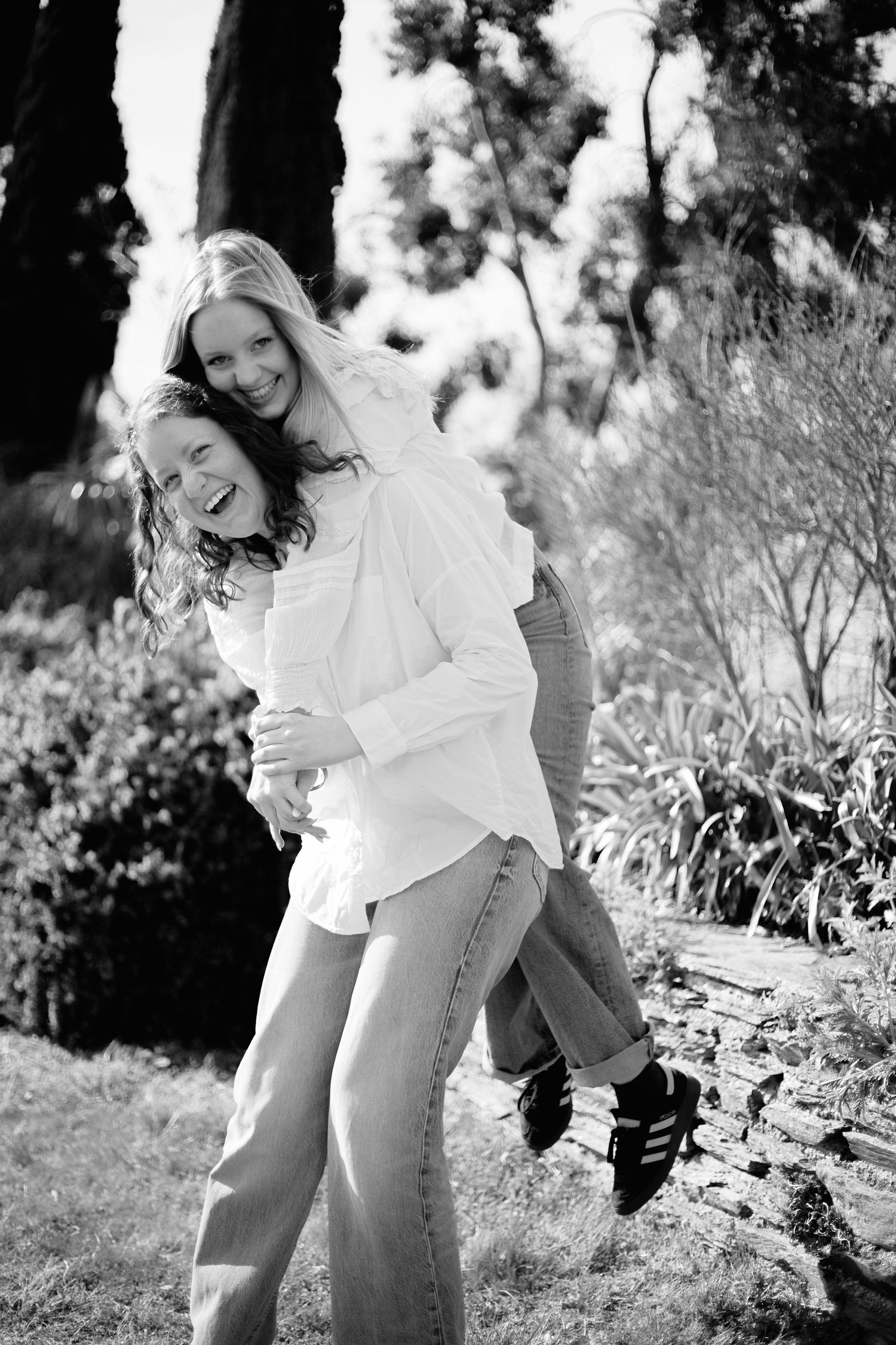 Two women laughing and sharing a piggyback ride outdoors in a garden, black and white photo.
