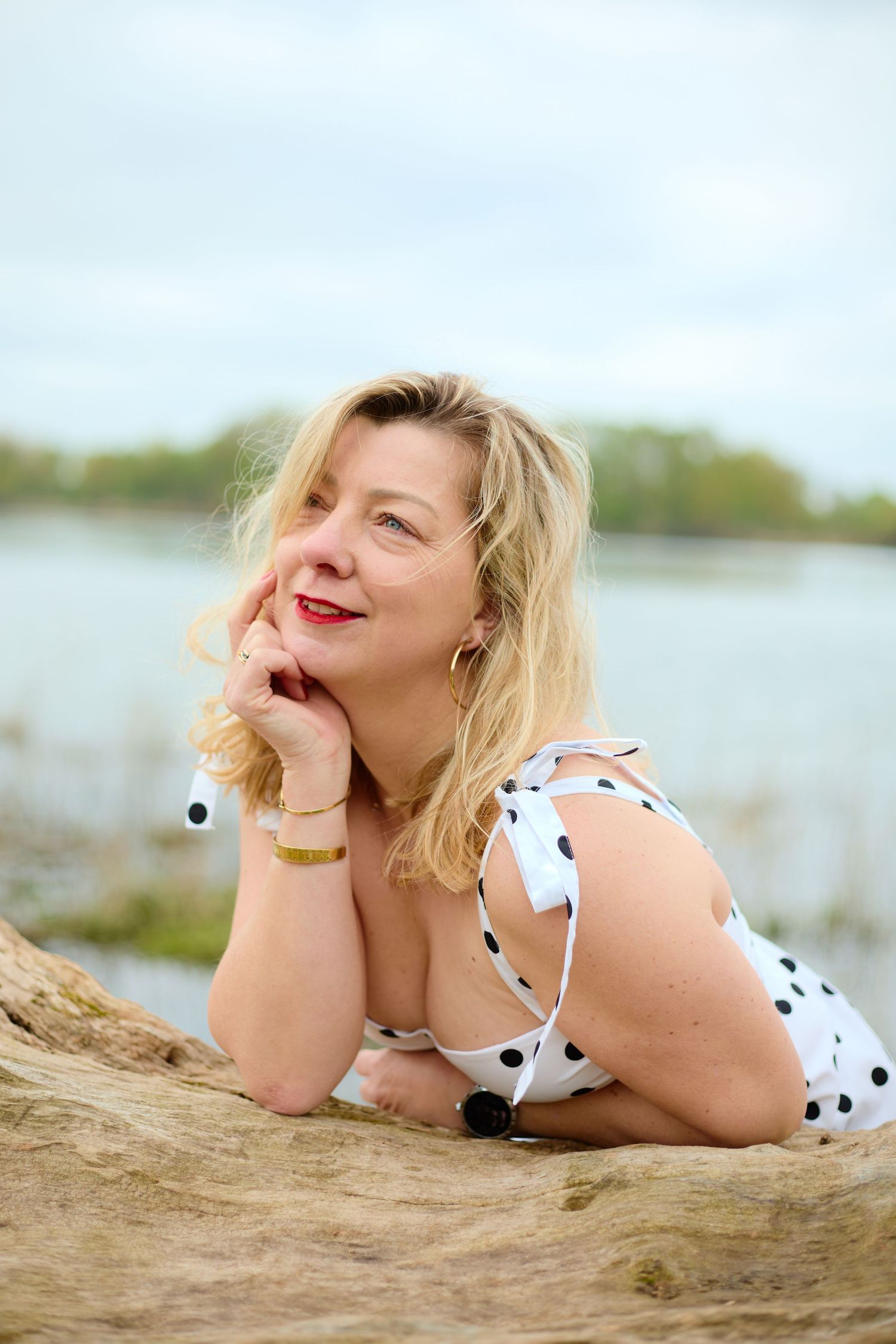 Blonde woman in white polka dot swimsuit posing on driftwood by a lake, resting chin on hand.