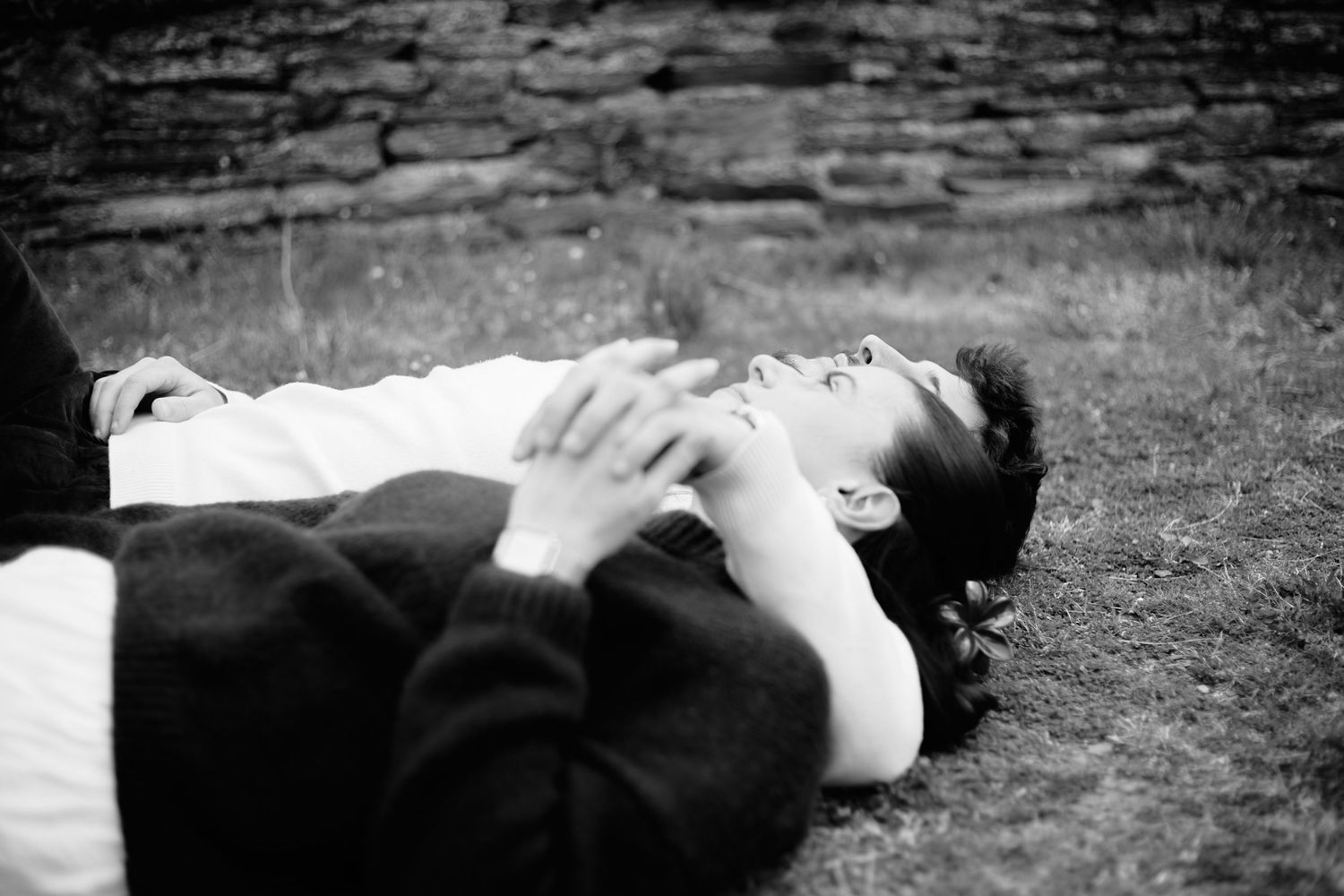 Black and white photo of a couple lying on grass, relaxing outdoors near a stone wall.