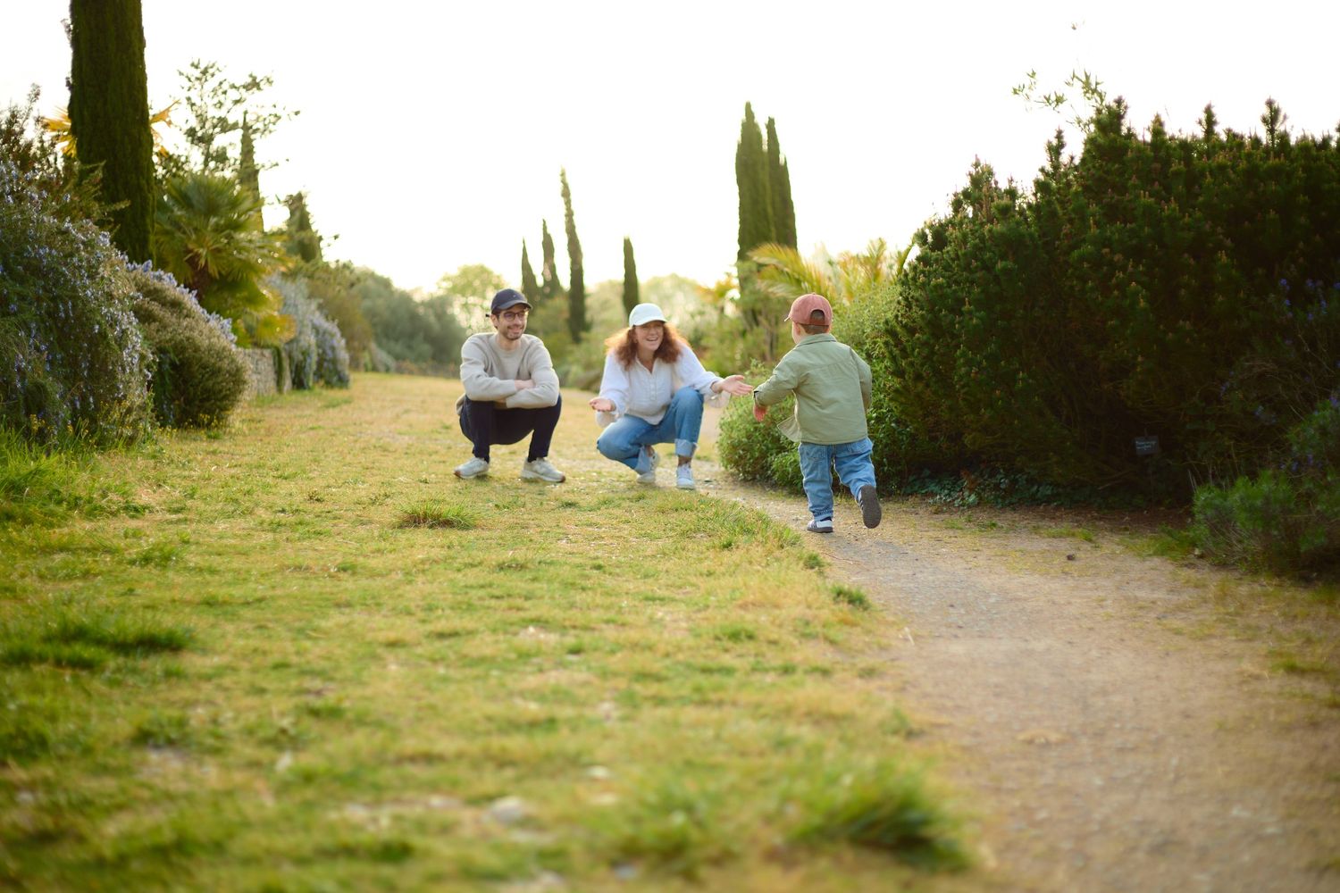Family playing outdoors on a sunny garden path surrounded by lush green hedges and cypress trees.