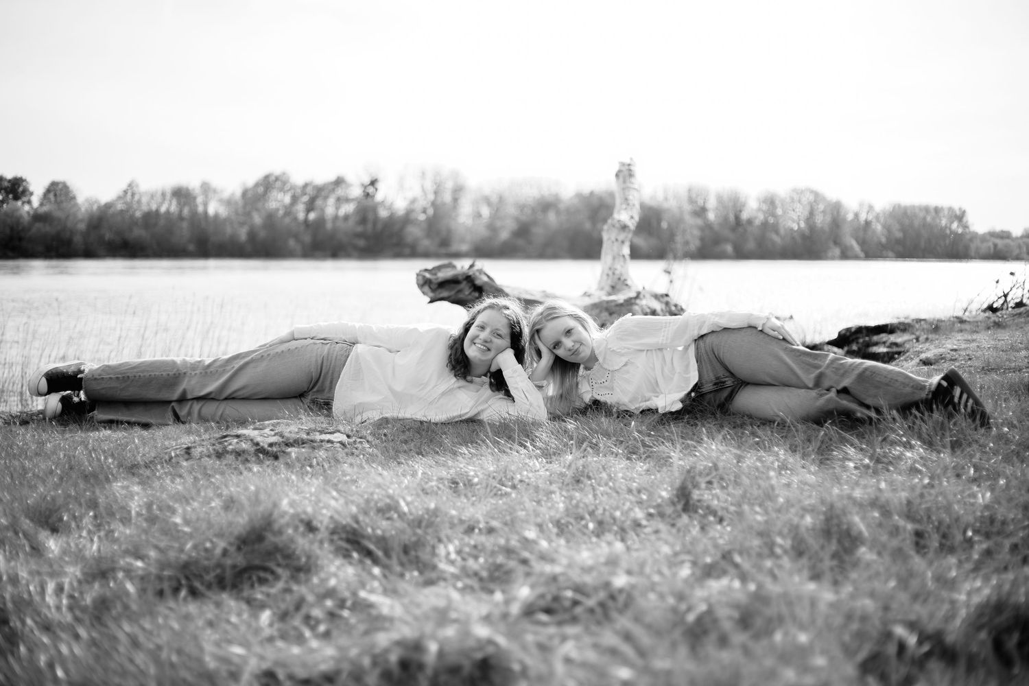 Black and white photo of two women lying in grass near a lake, laughing together on a sunny day.