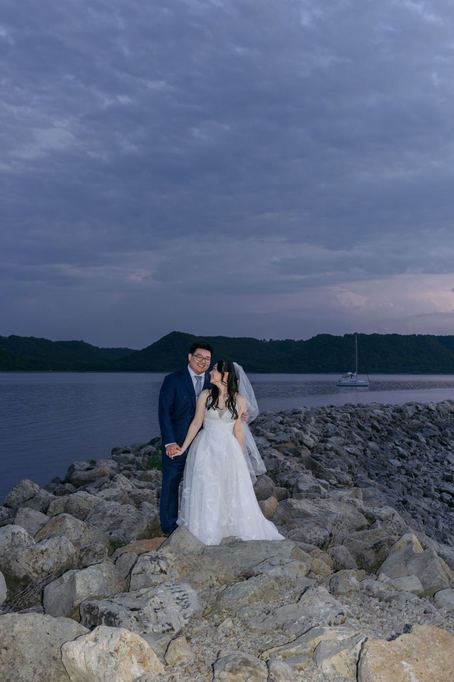 Bride and groom pose on rocky shoreline at dusk, with serene water and cloudy sky backdrop.