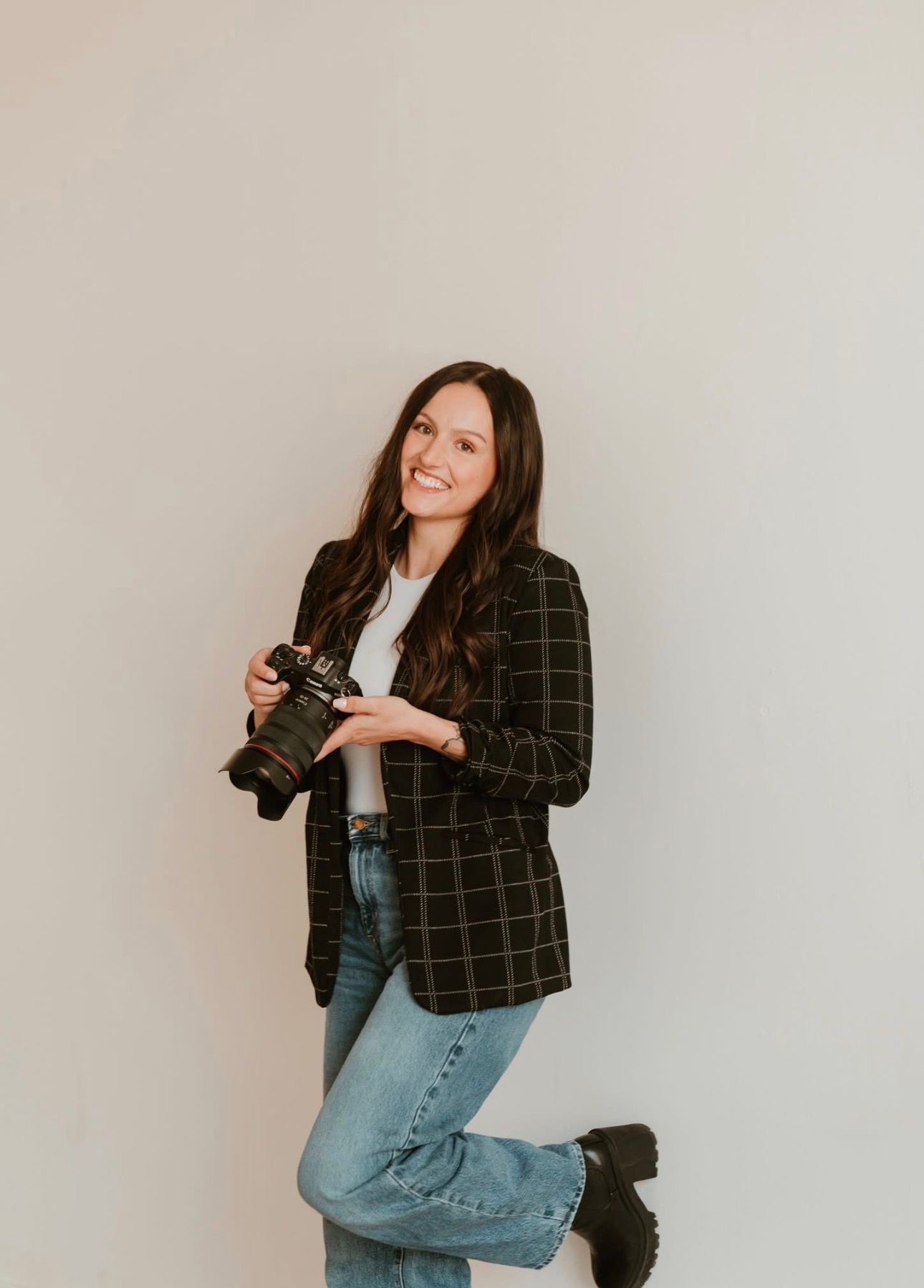 Smiling brunette woman holding a camera wearing a plaid blazer and jeans posing against a white wall.