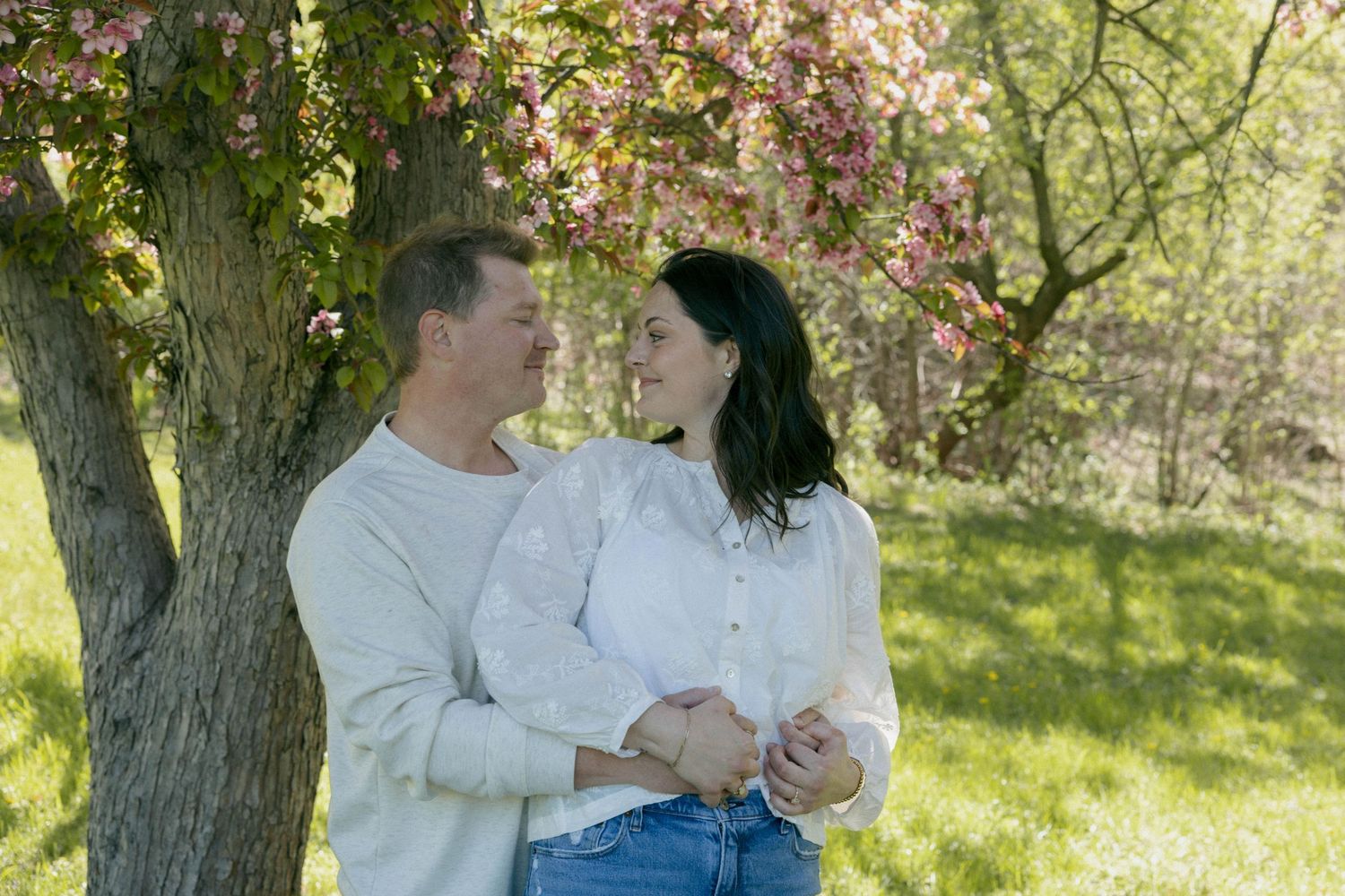 A couple smiles at each other while holding hands under a blooming pink tree in a lush green garden.