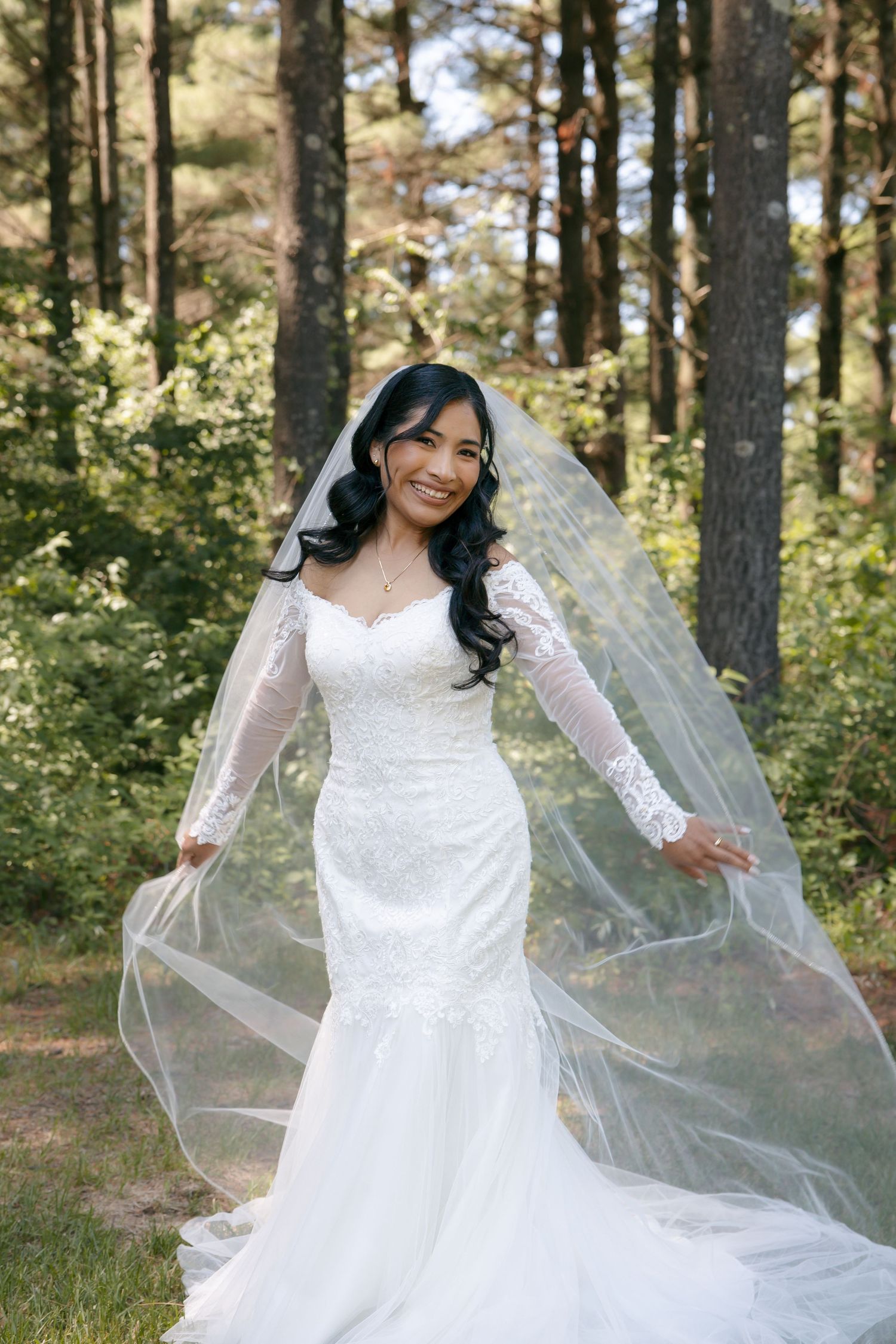 Smiling bride in white lace mermaid gown with long veil poses outdoors in a pine forest on her wedding day.
