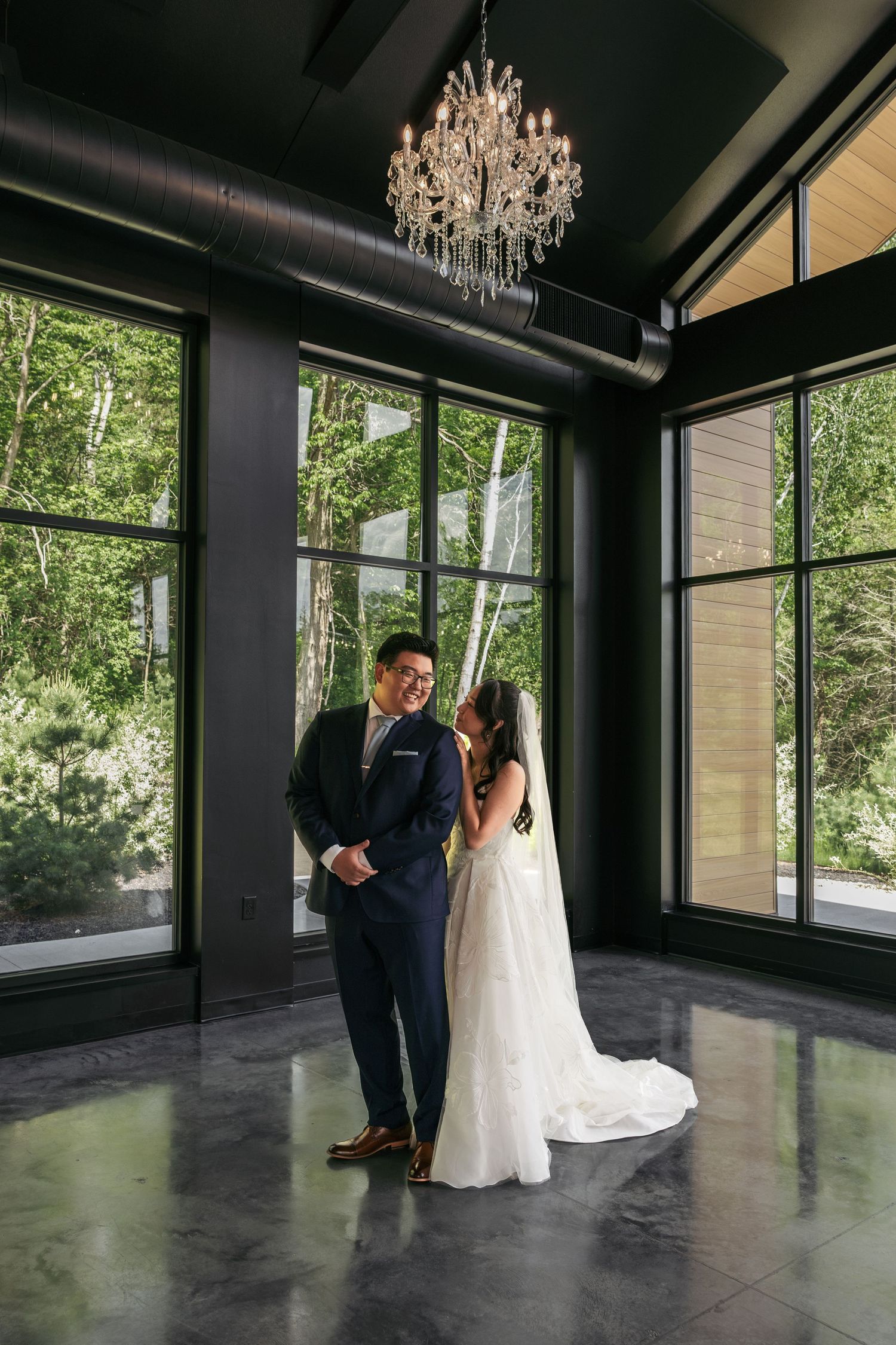 Bride and groom share an intimate moment under a crystal chandelier in a modern venue with floor-to-ceiling windows.