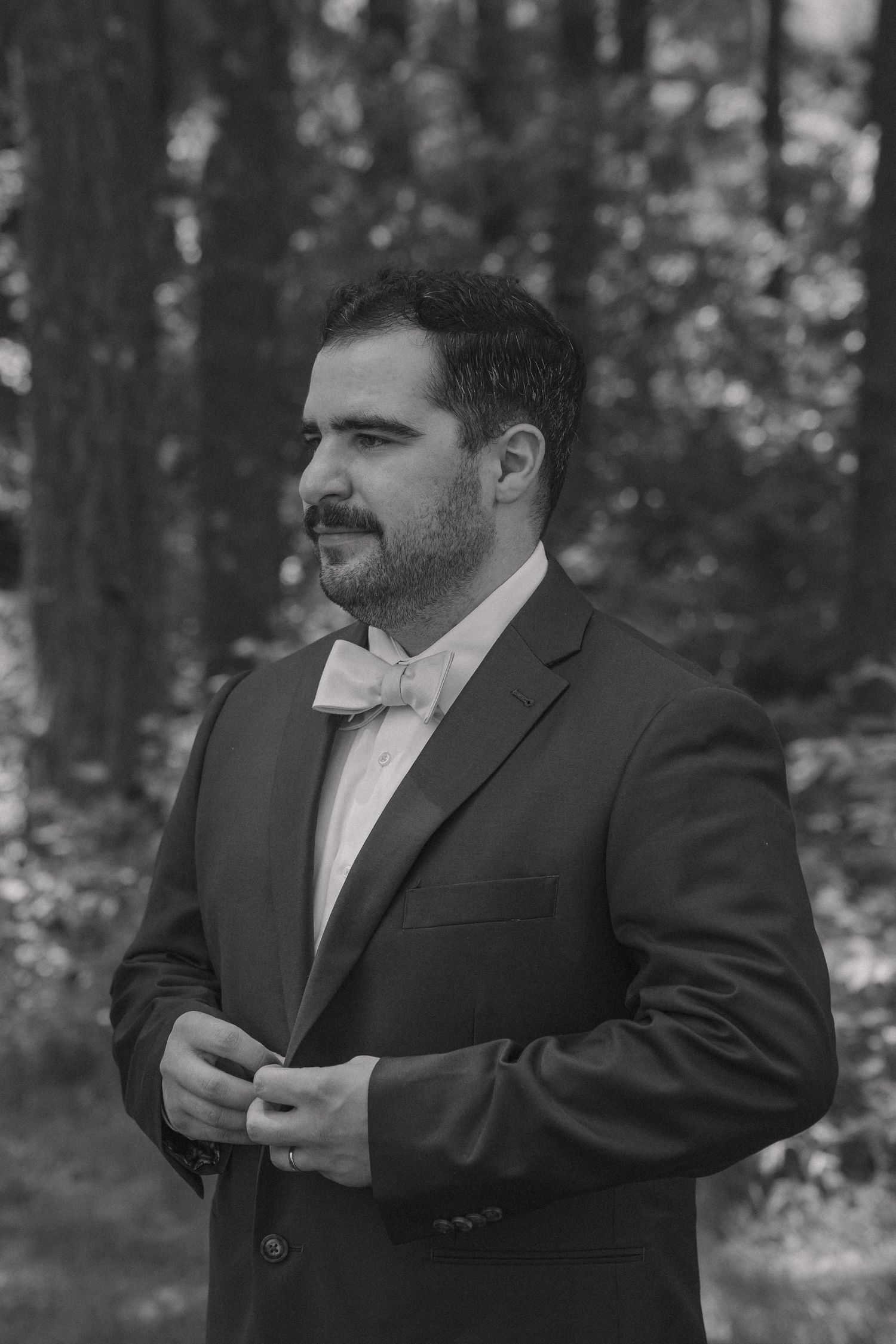 Black and white portrait of groom in suit and bow tie standing among tall trees.