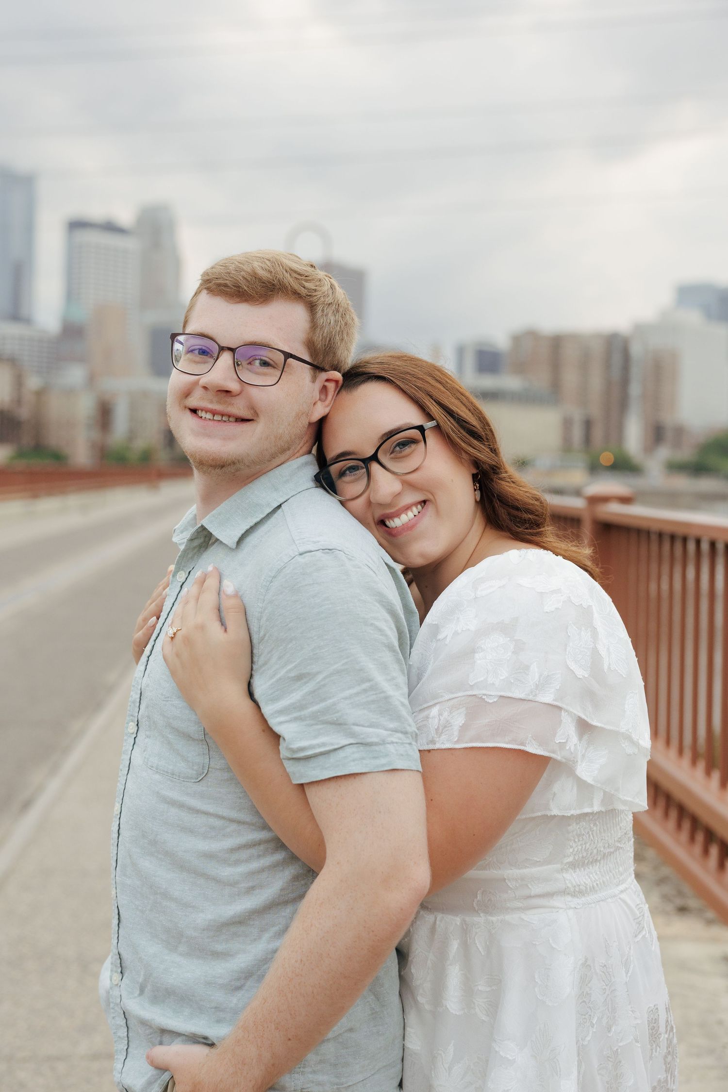 Happy couple posing on a bridge, woman leaning on mans shoulder with city skyline behind them.