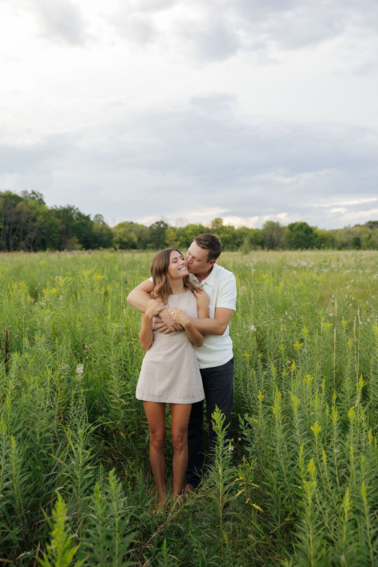 Man kissing woman on the cheek while embracing her from behind in a green meadow field.