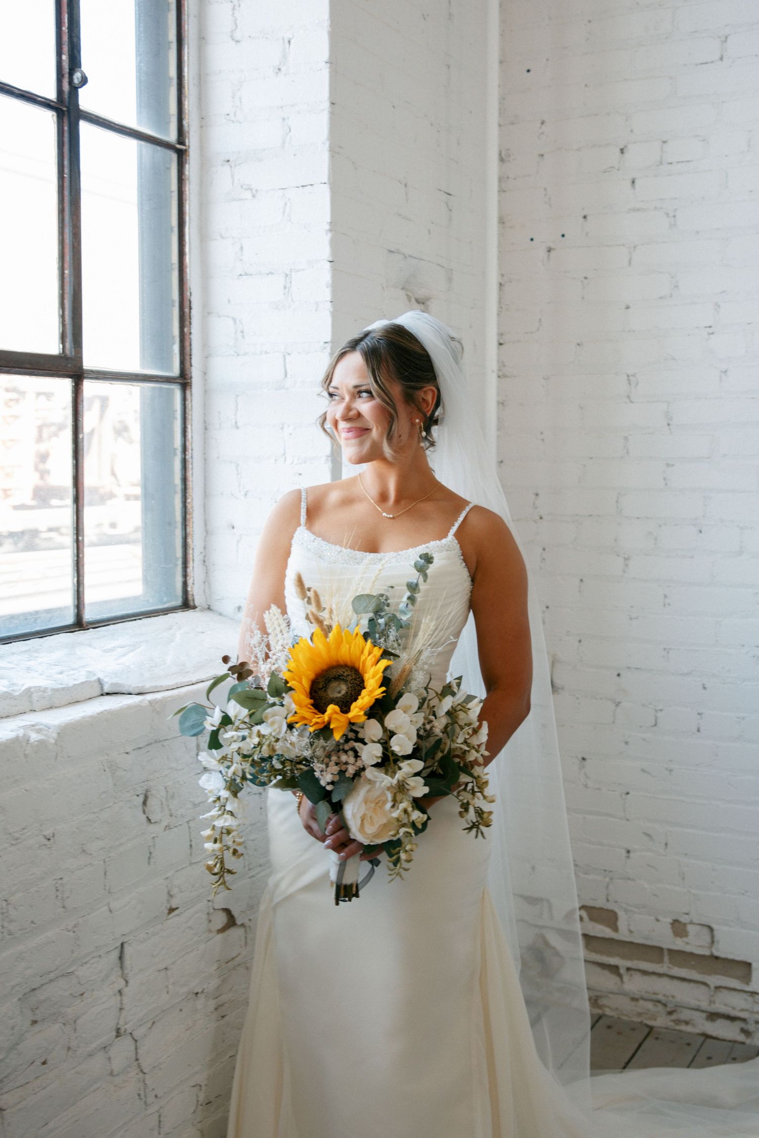 Smiling bride in white spaghetti strap gown holds sunflower bouquet by window in white brick venue.