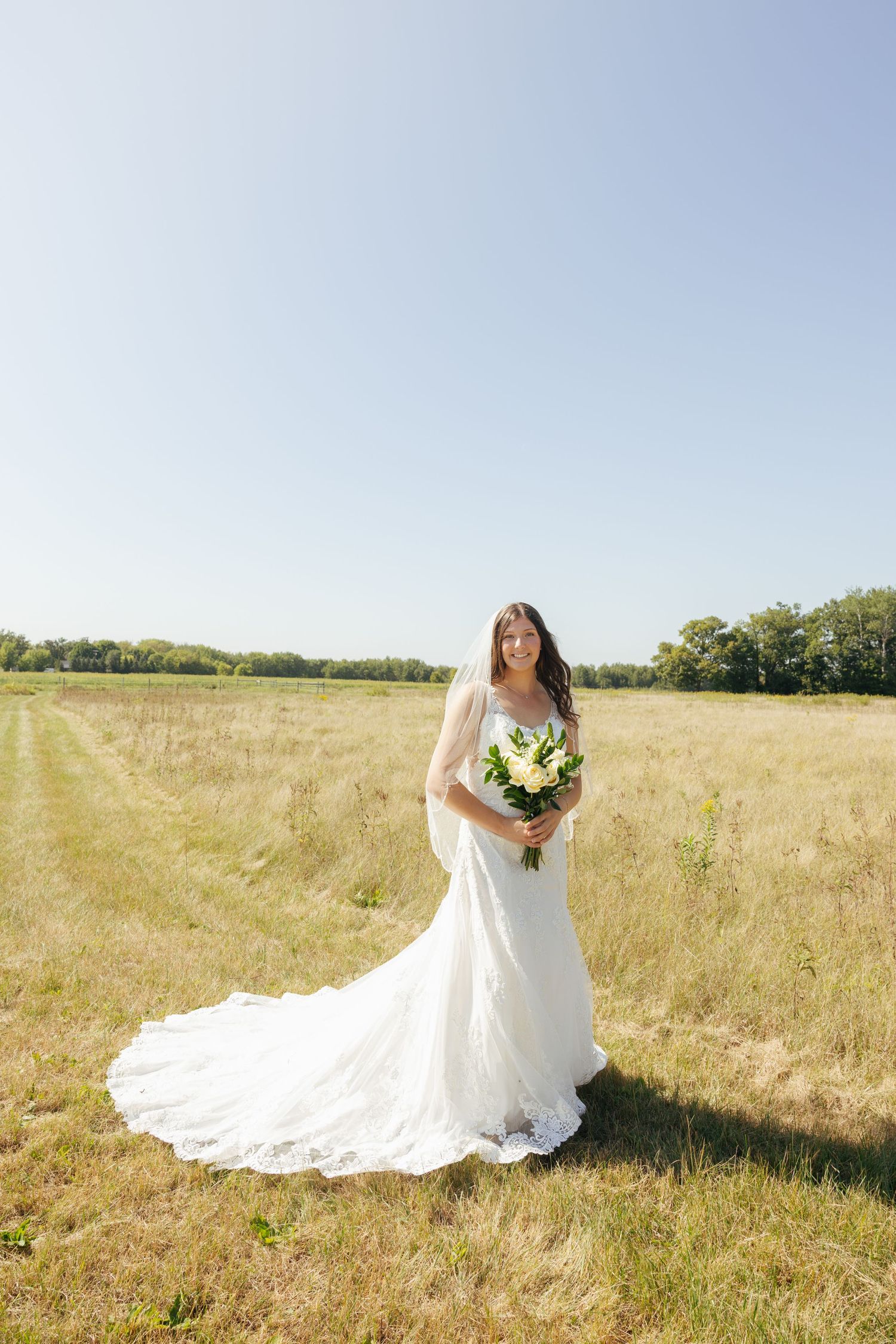 Bride in white lace gown with train holds bouquet in sunny green field.