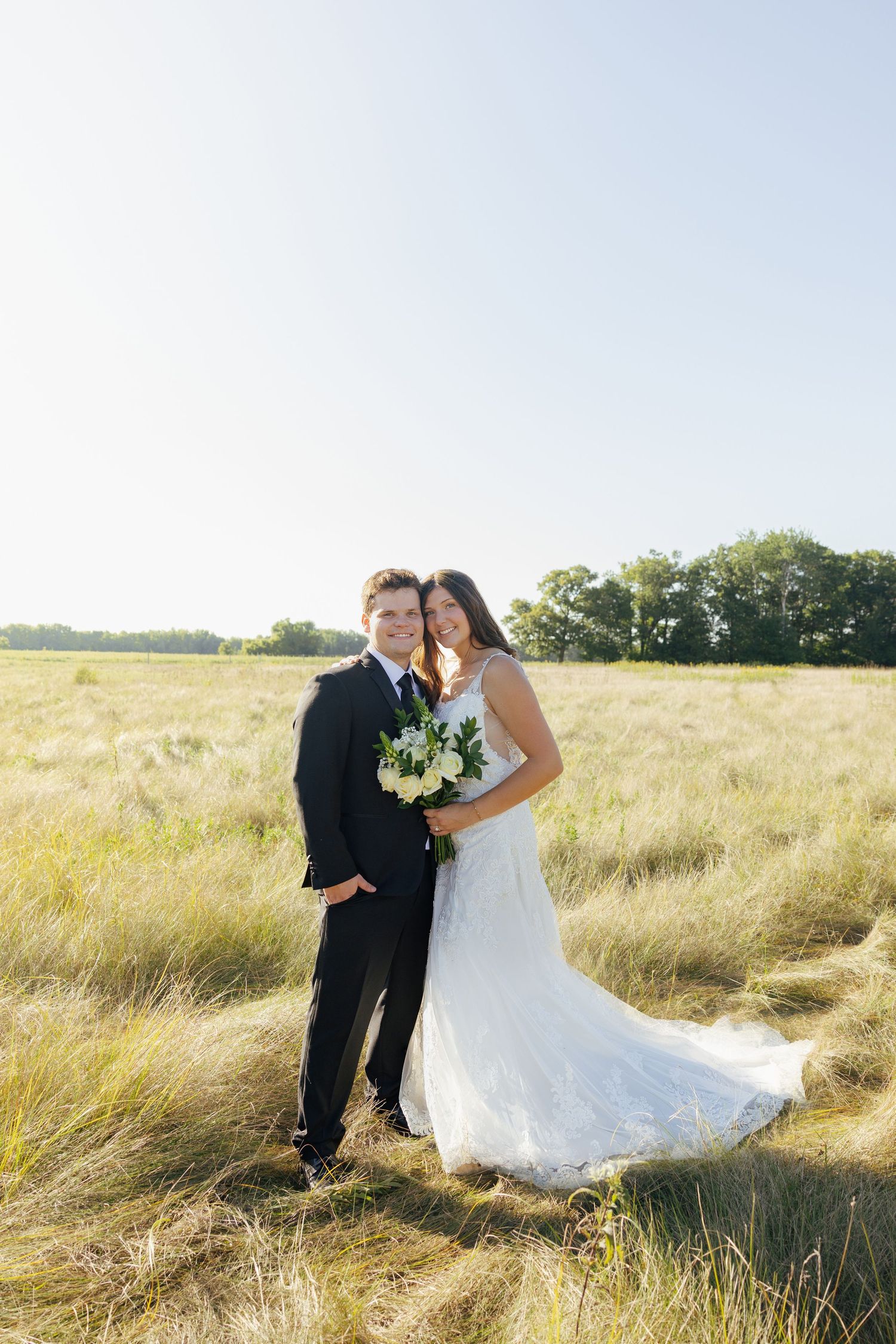 Smiling bride and groom posing together in a sunny golden grass field on their wedding day.