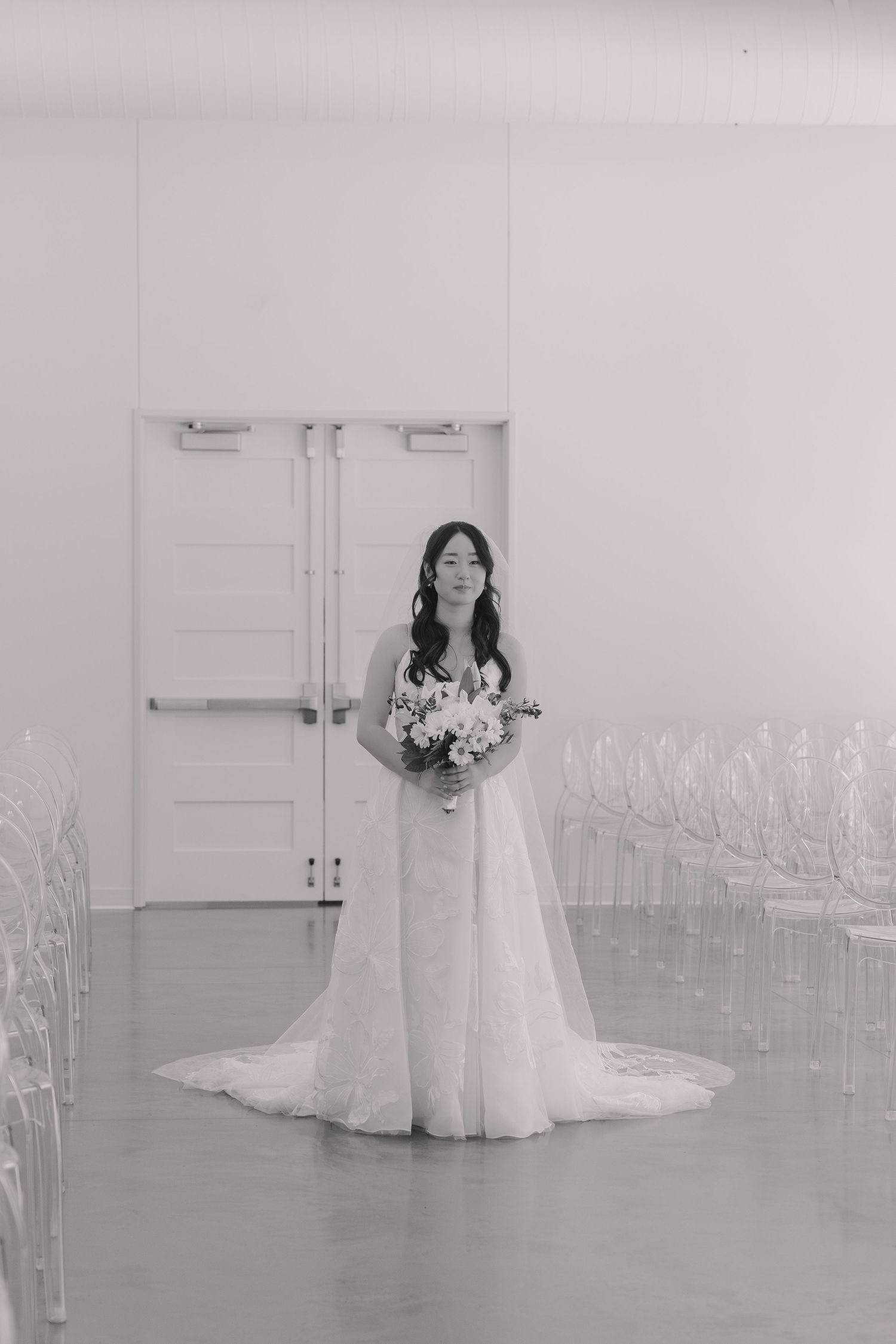 Bride in lace wedding gown holding floral bouquet stands in white ceremony venue with ghost chairs.