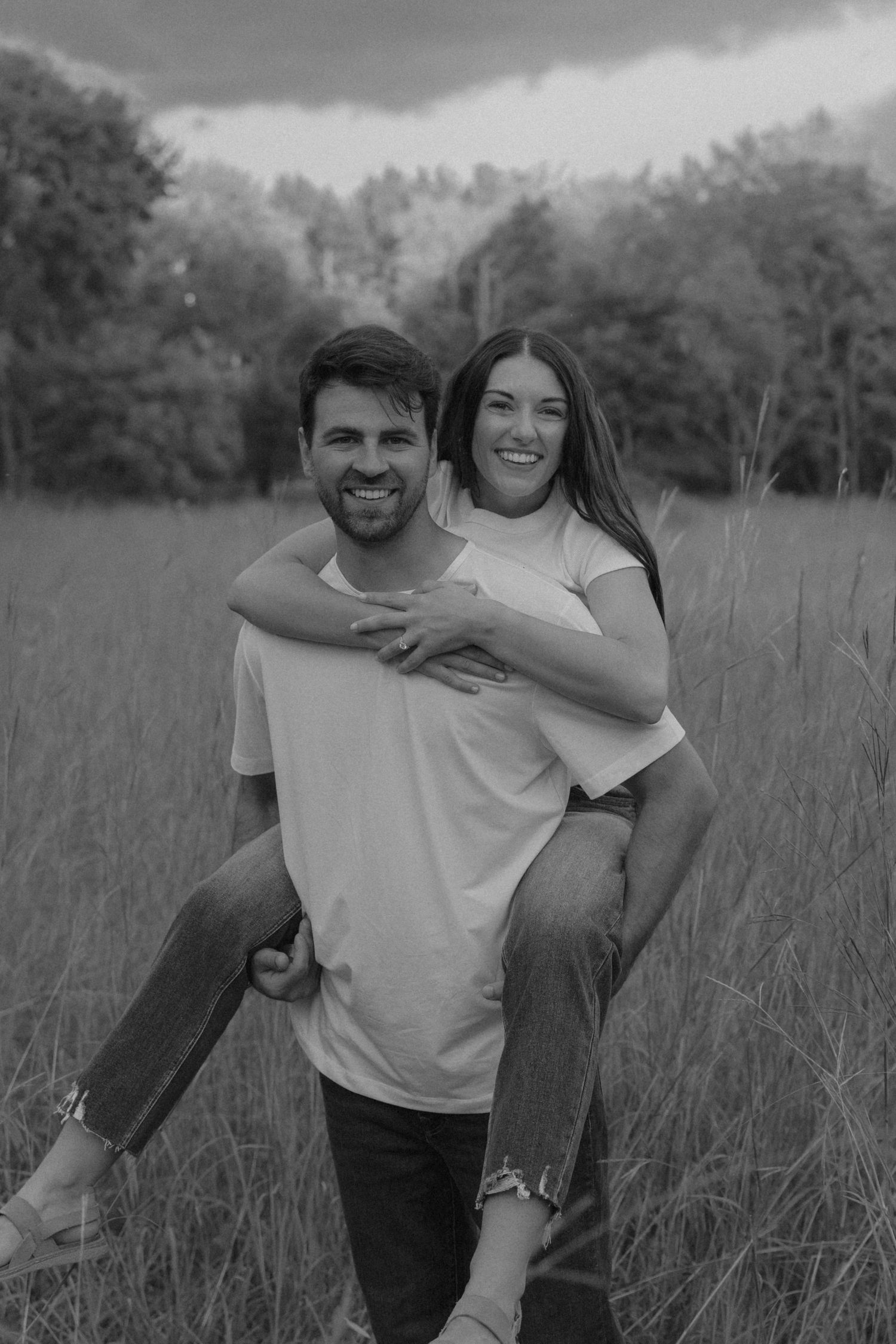 Black and white photo of a smiling couple in a field, man giving woman a piggyback ride outdoors.