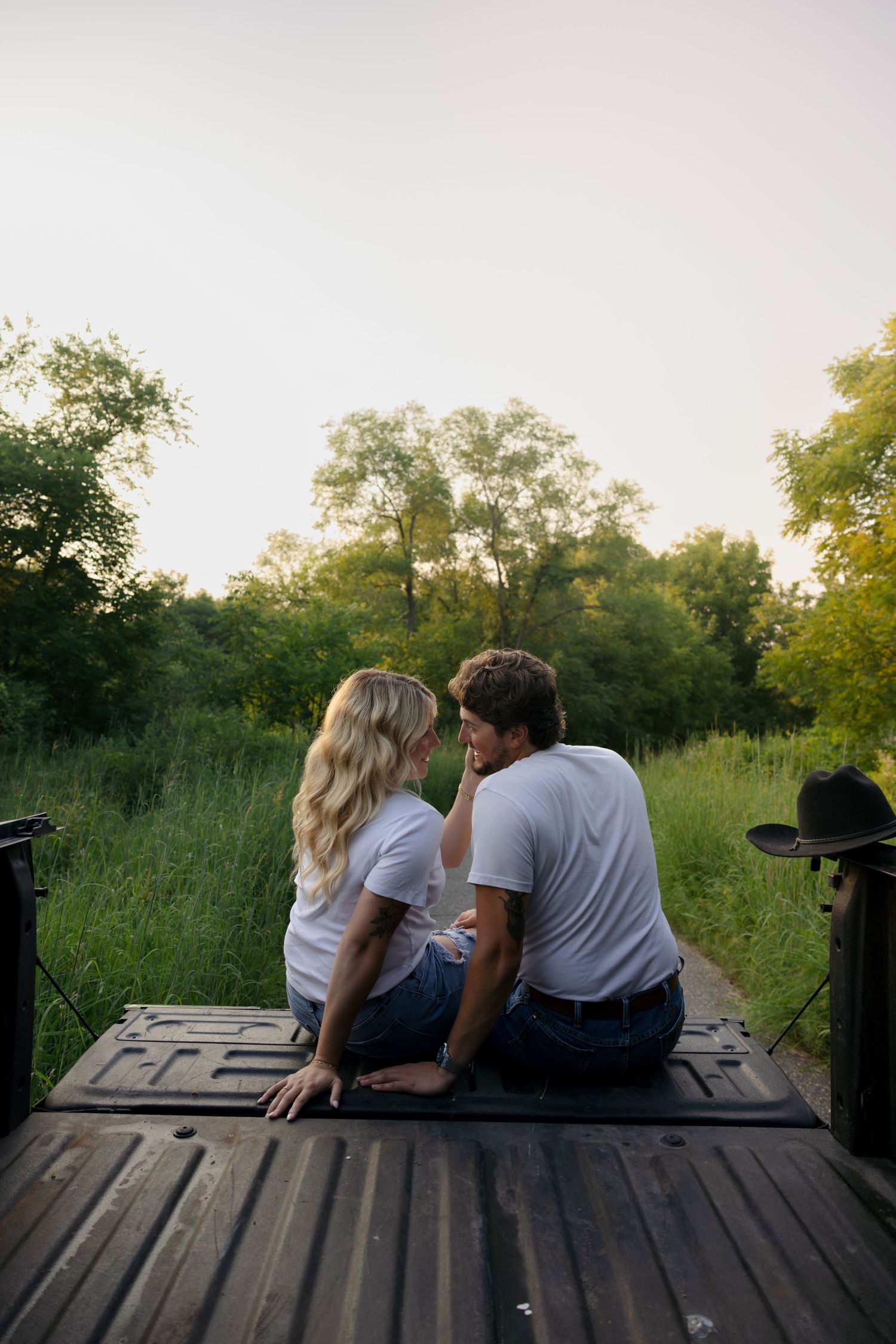 Couple sitting closely on a truck bed tailgate, facing each other in a lush green field at dusk.