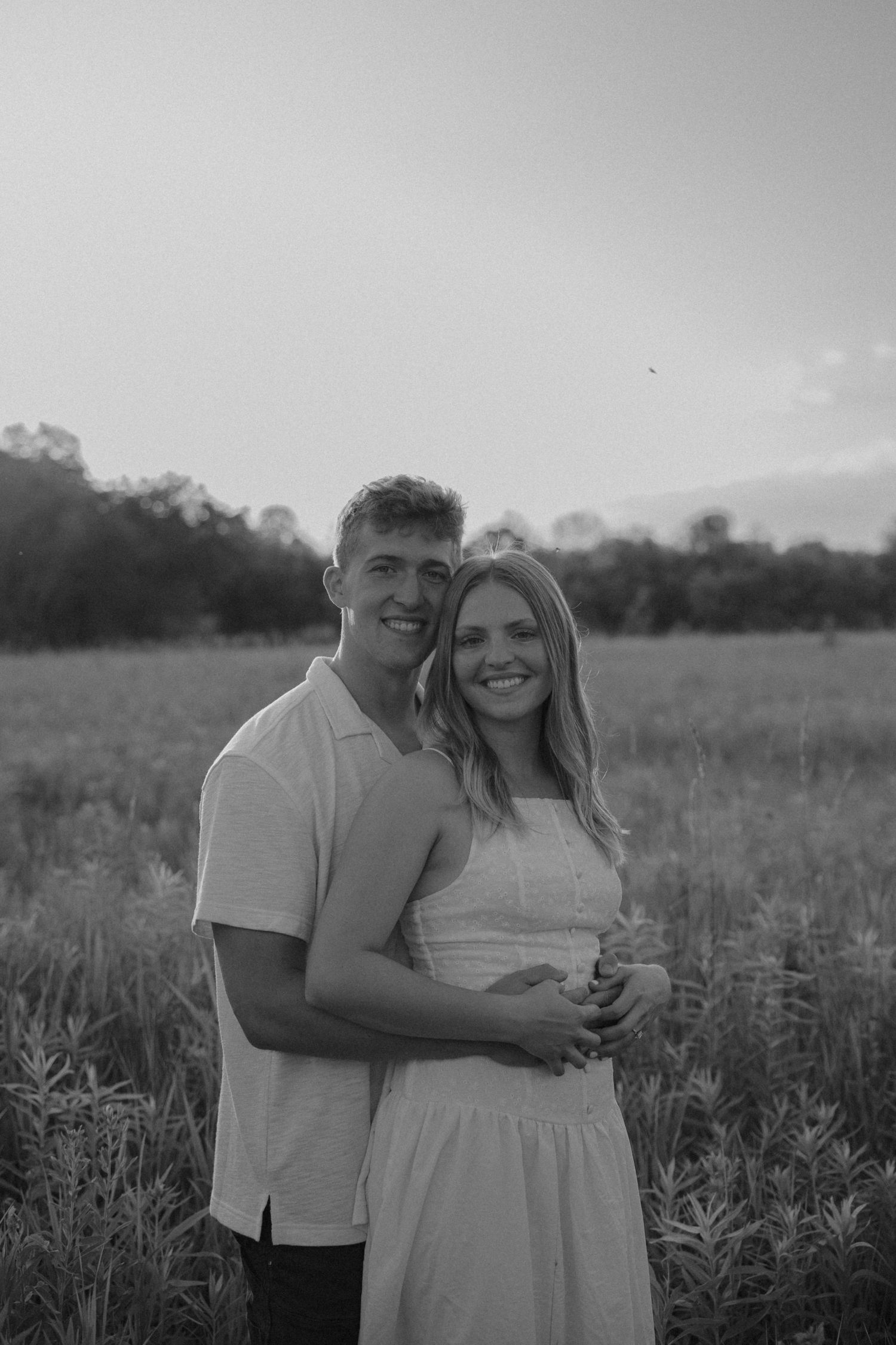 Black and white photo of a smiling couple embracing in a wildflower field at sunset.