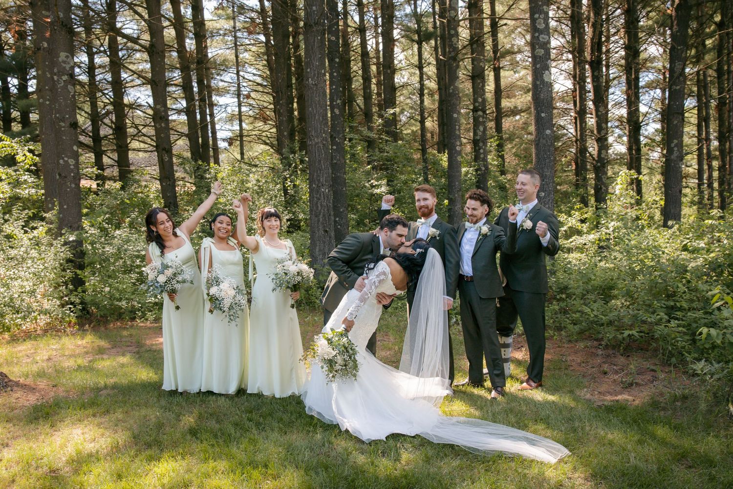 Wedding party celebrates in a forest clearing, bride and groom kissing while bridesmaids and groomsmen cheer around them.