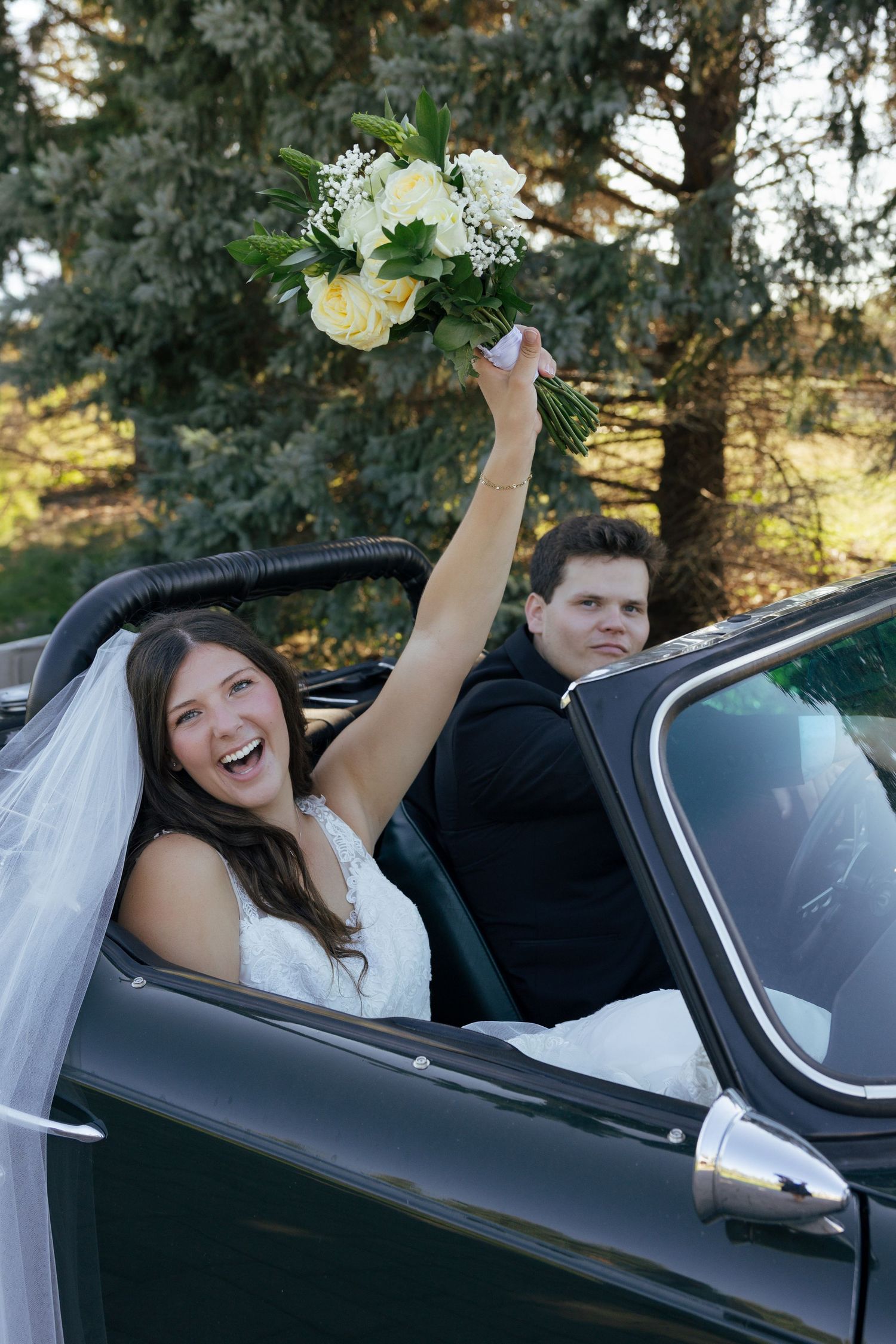 Joyful bride raising white rose bouquet in a convertible car with groom seated beside her on wedding day.