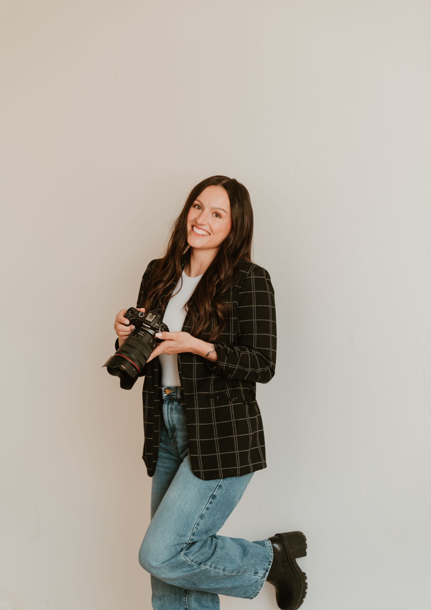 Smiling brunette woman holding a DSLR camera, wearing a plaid blazer and jeans, posing against a light gray background.