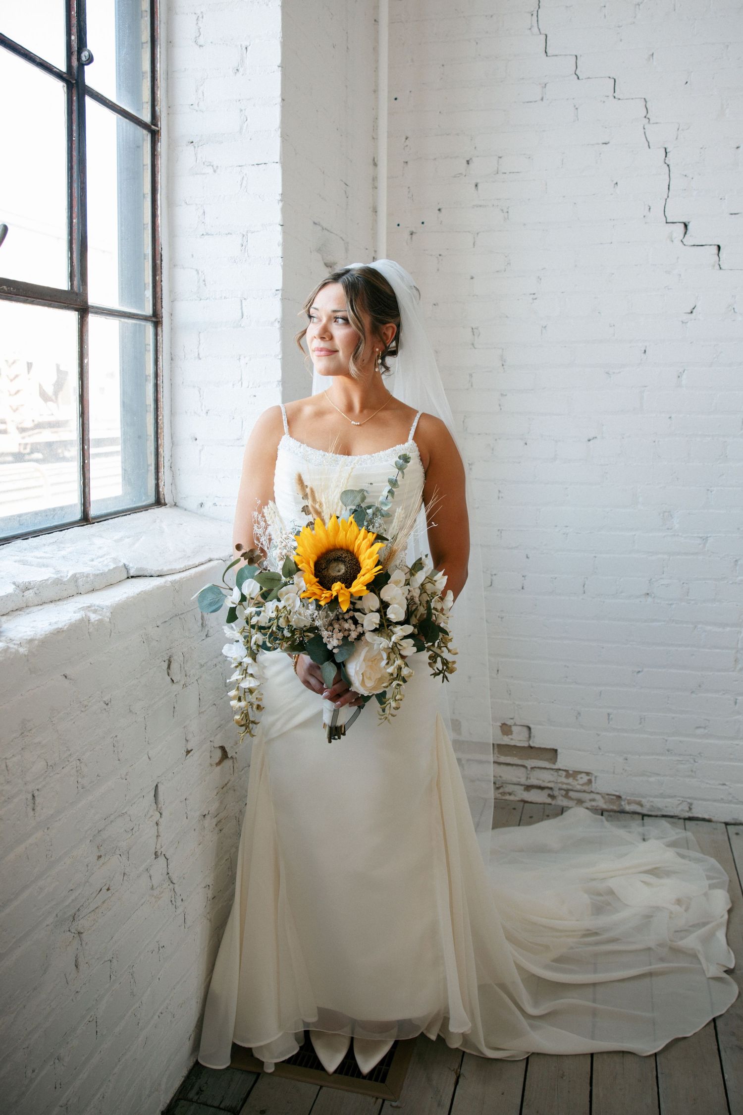 Bride in white gown holding sunflower bouquet standing by large window in white brick room.