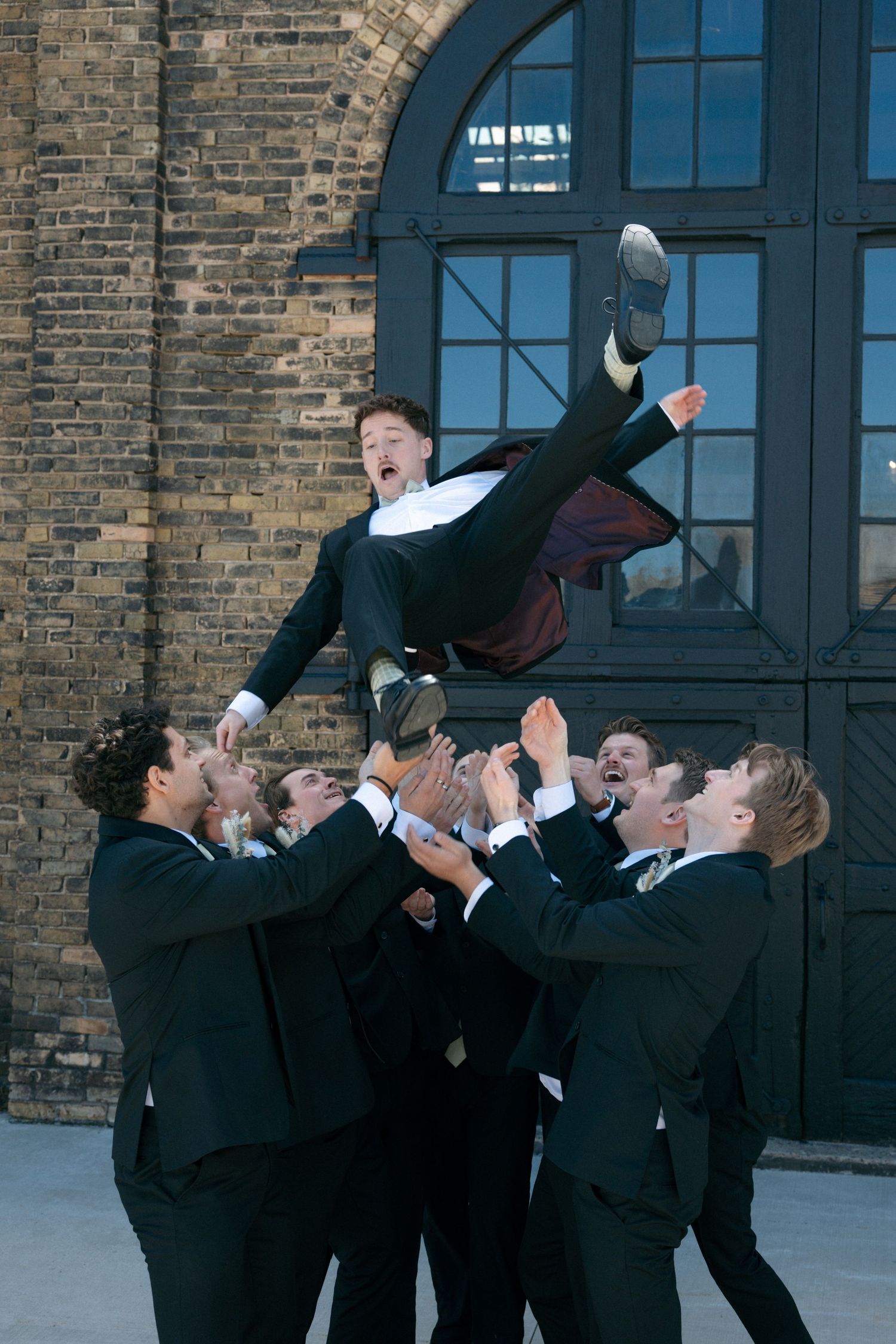 Groomsmen tossing the groom into the air outside a brick building during wedding celebrations.
