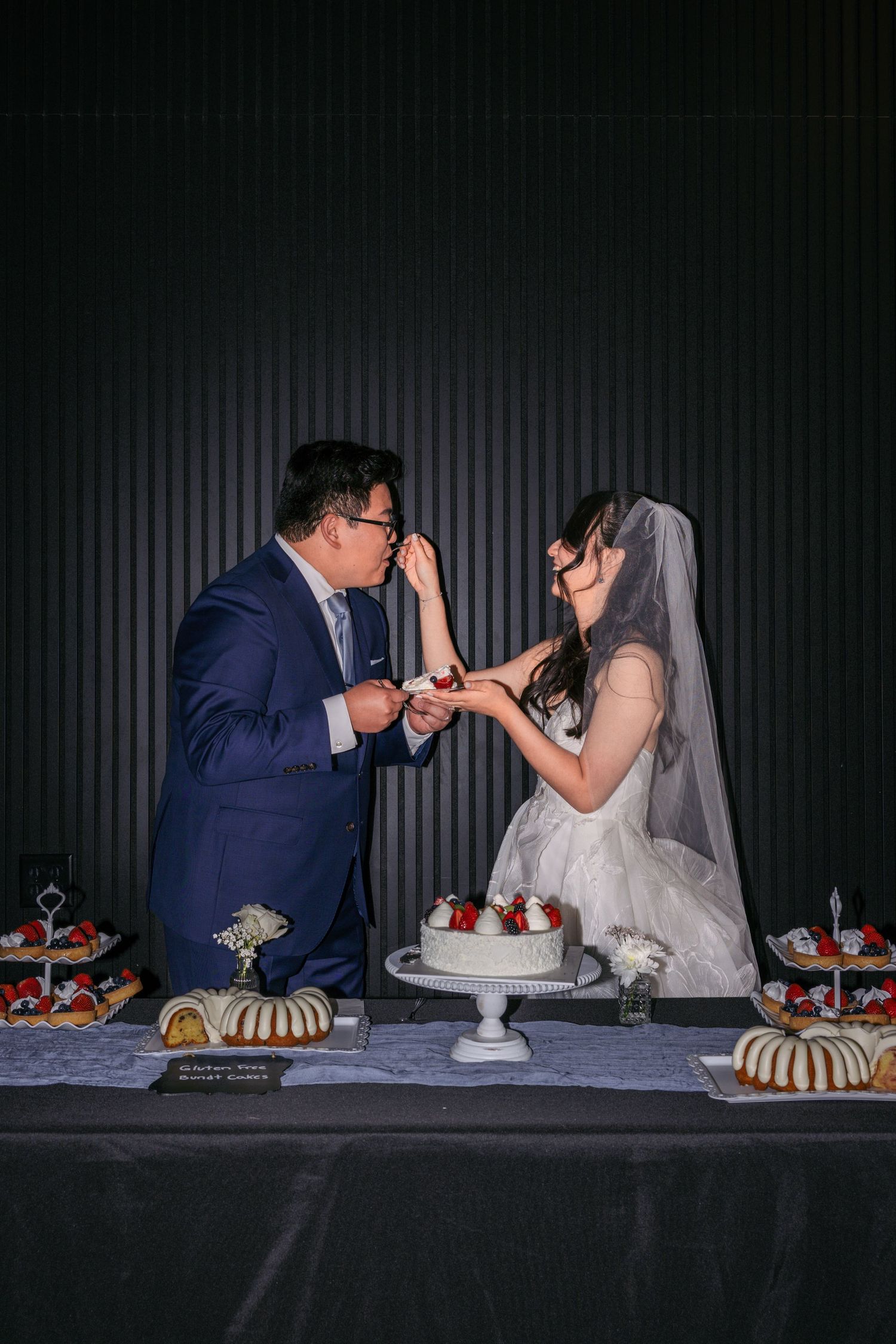 Bride and groom feed each other cake at a wedding dessert table with a white tiered cake and strawberries.