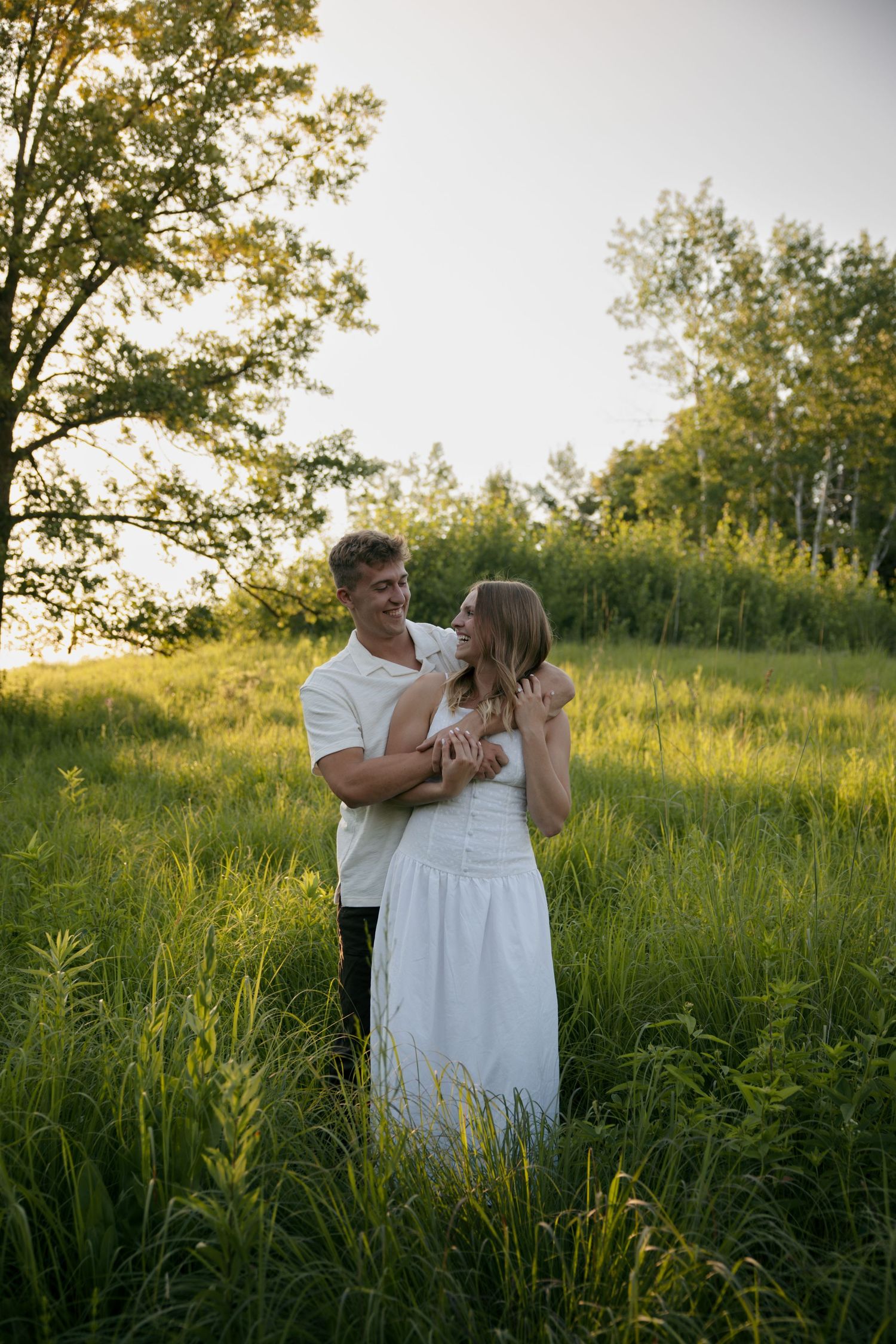 Couple holding each other in lush green field at golden hour, gazing lovingly into each other's eyes.
