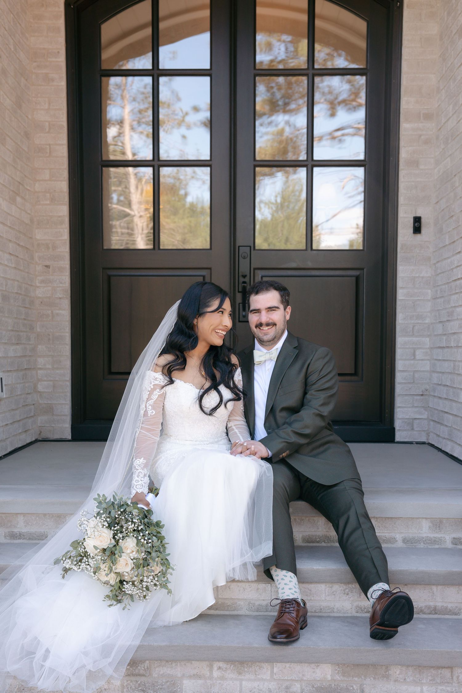 Bride and groom sitting on steps before dark double doors, bride holding white bouquet in elegant lace wedding gown.