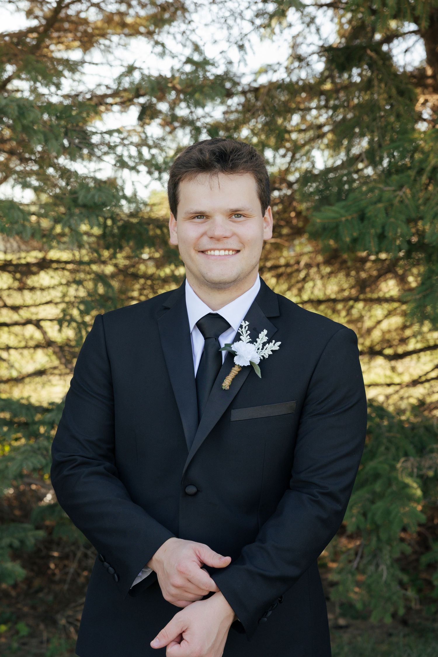 Smiling groom in black suit with white boutonniere posing outdoors among green trees.