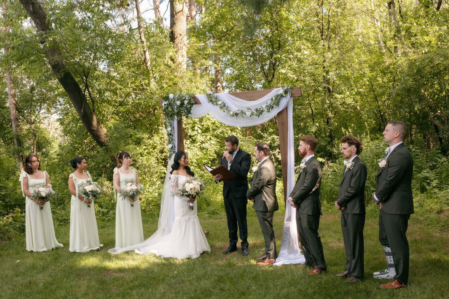 Outdoor wedding ceremony under floral arch with bridesmaids in white, groomsmen in green suits, and officiant in lush garden.