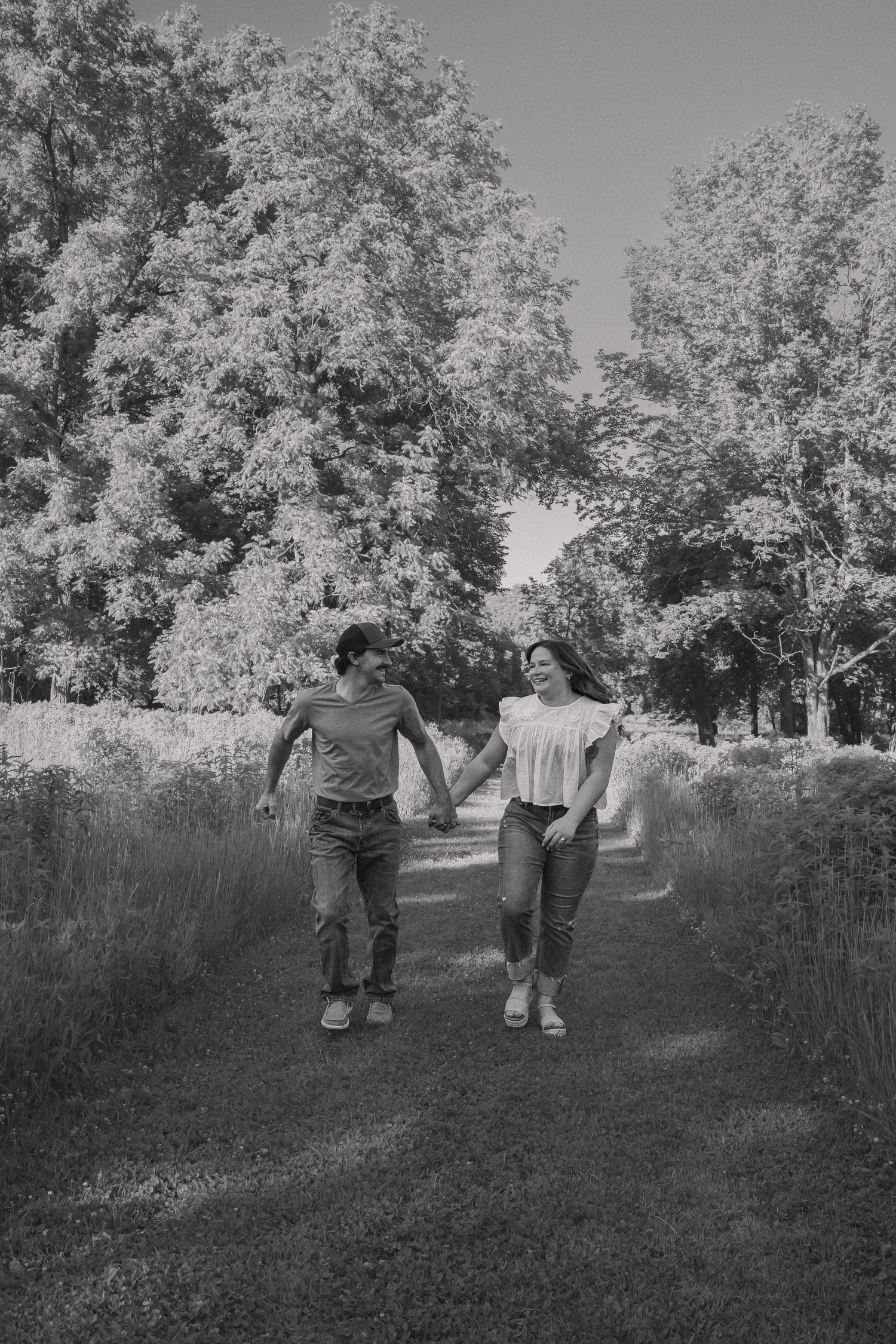 Black and white photo of a couple holding hands and walking together on a scenic tree-lined path outdoors.