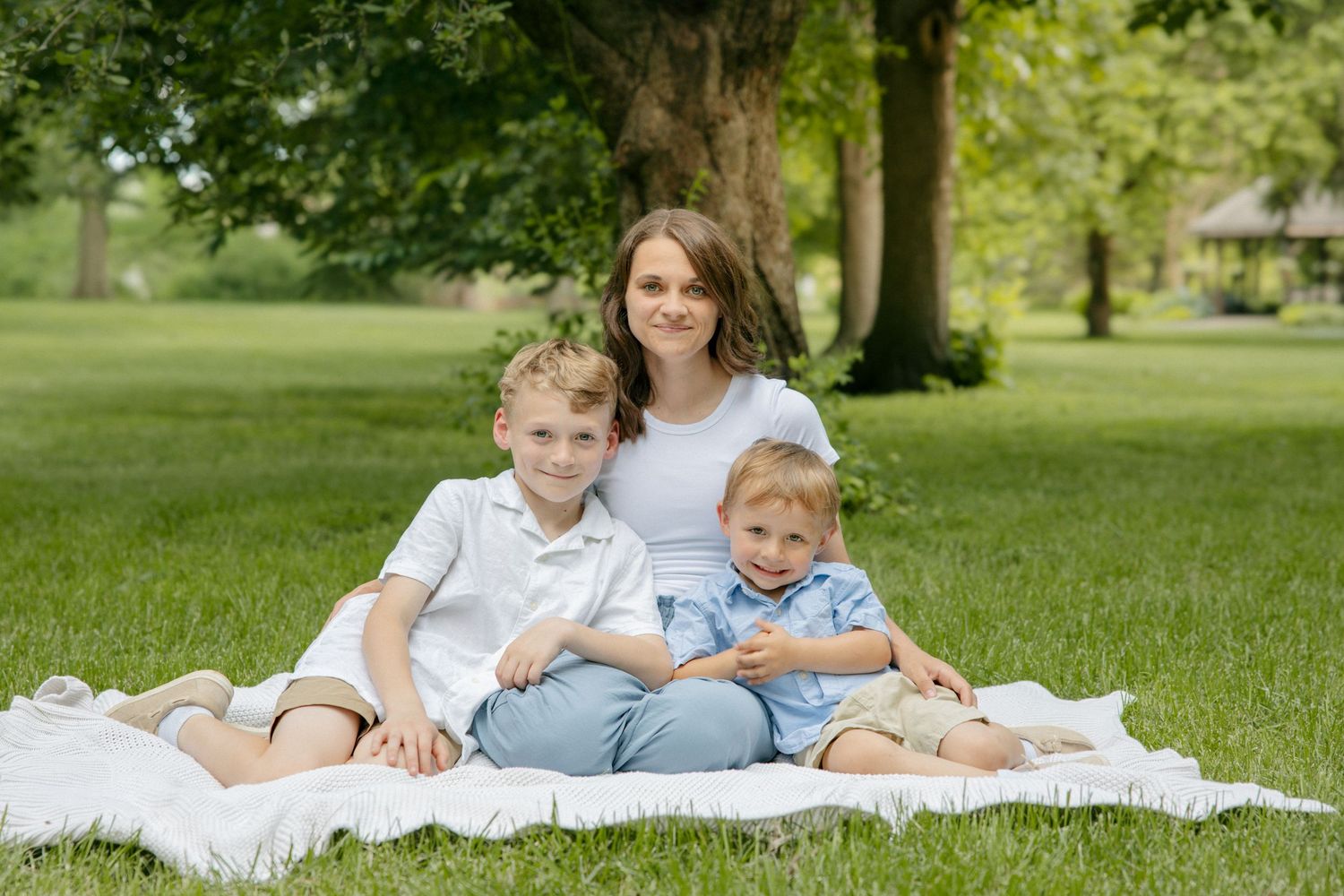 A mother sits on a white blanket in a park with her two young sons under a large green tree.