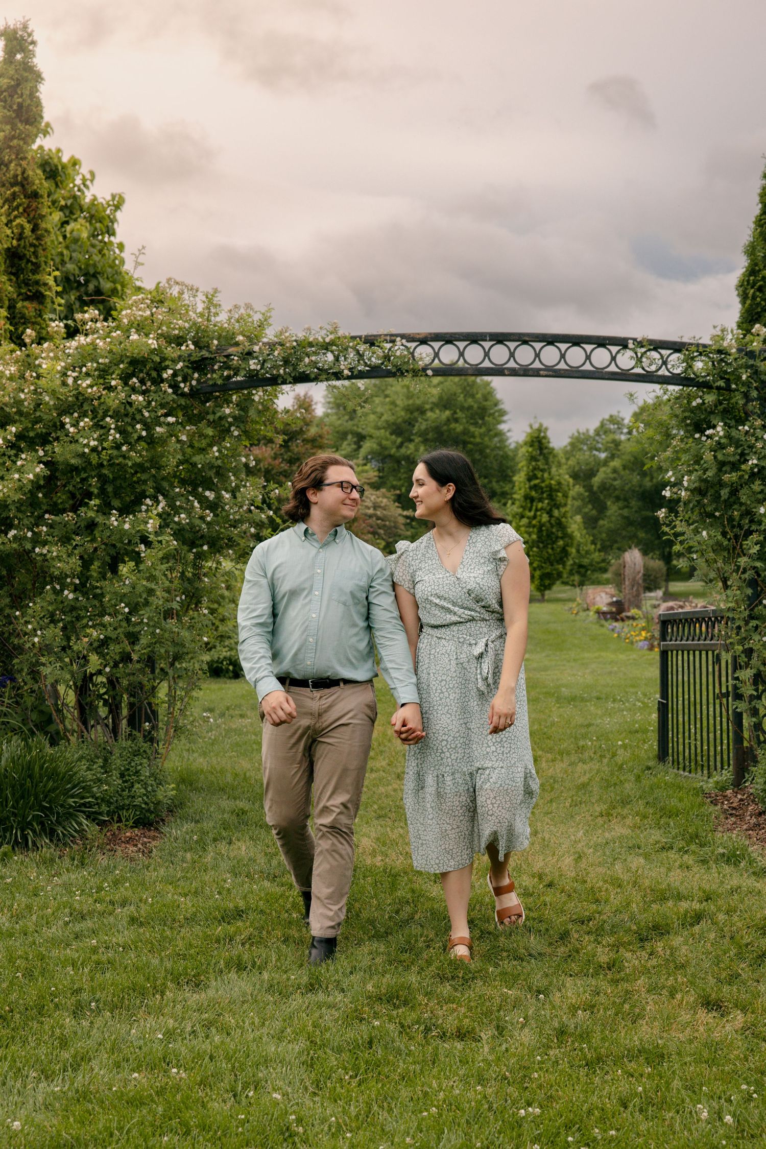 A couple walks hand-in-hand through a lush garden arch, smiling at each other on a cloudy day.