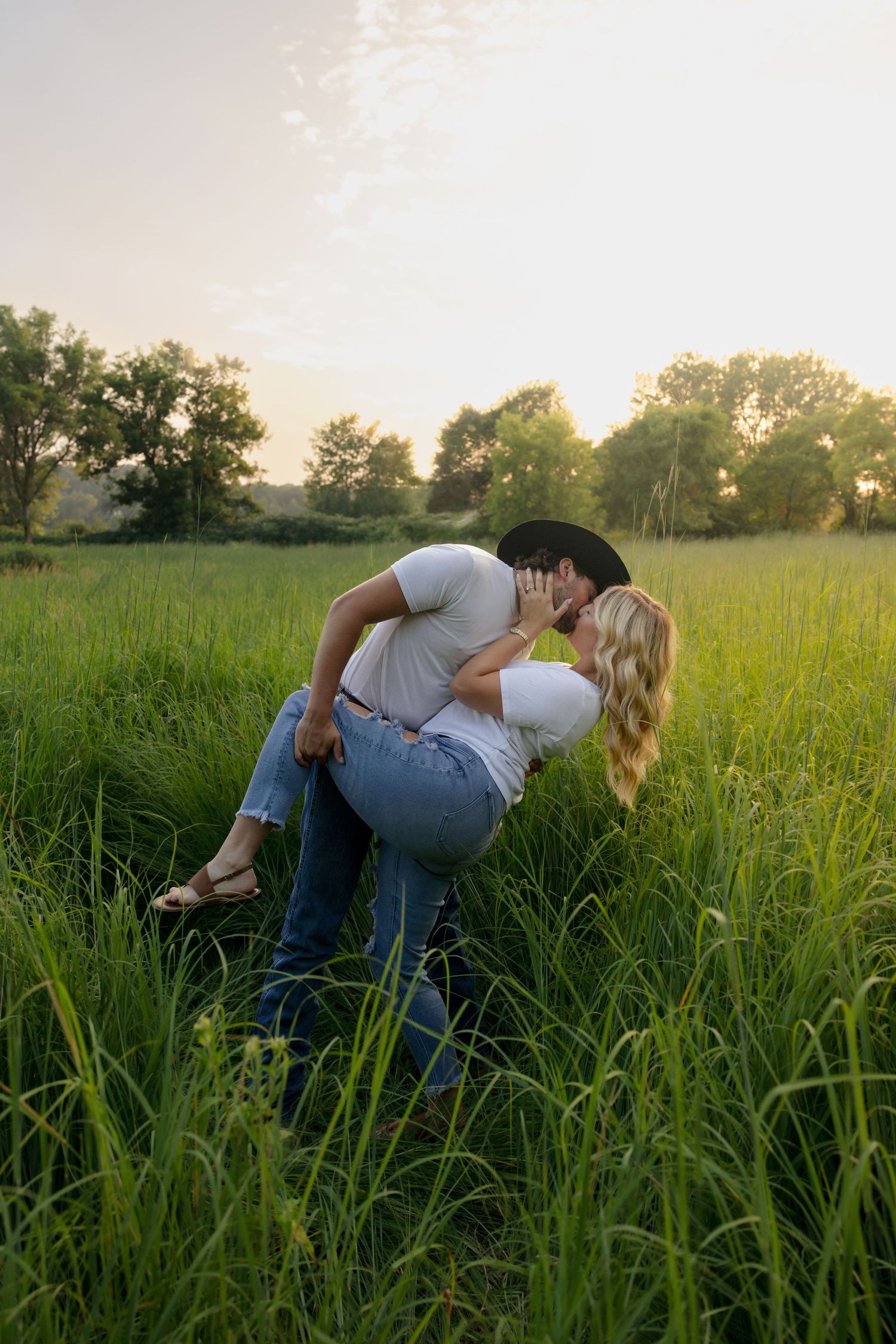 Romantic couple kissing in lush green field at sunset, man dipping woman in jeans and white tees.