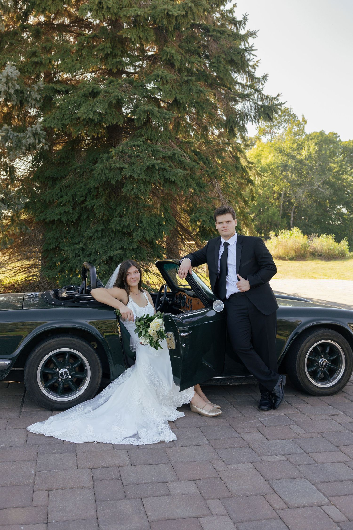 Bride and groom posing elegantly beside a classic black convertible car outdoors on their wedding day.