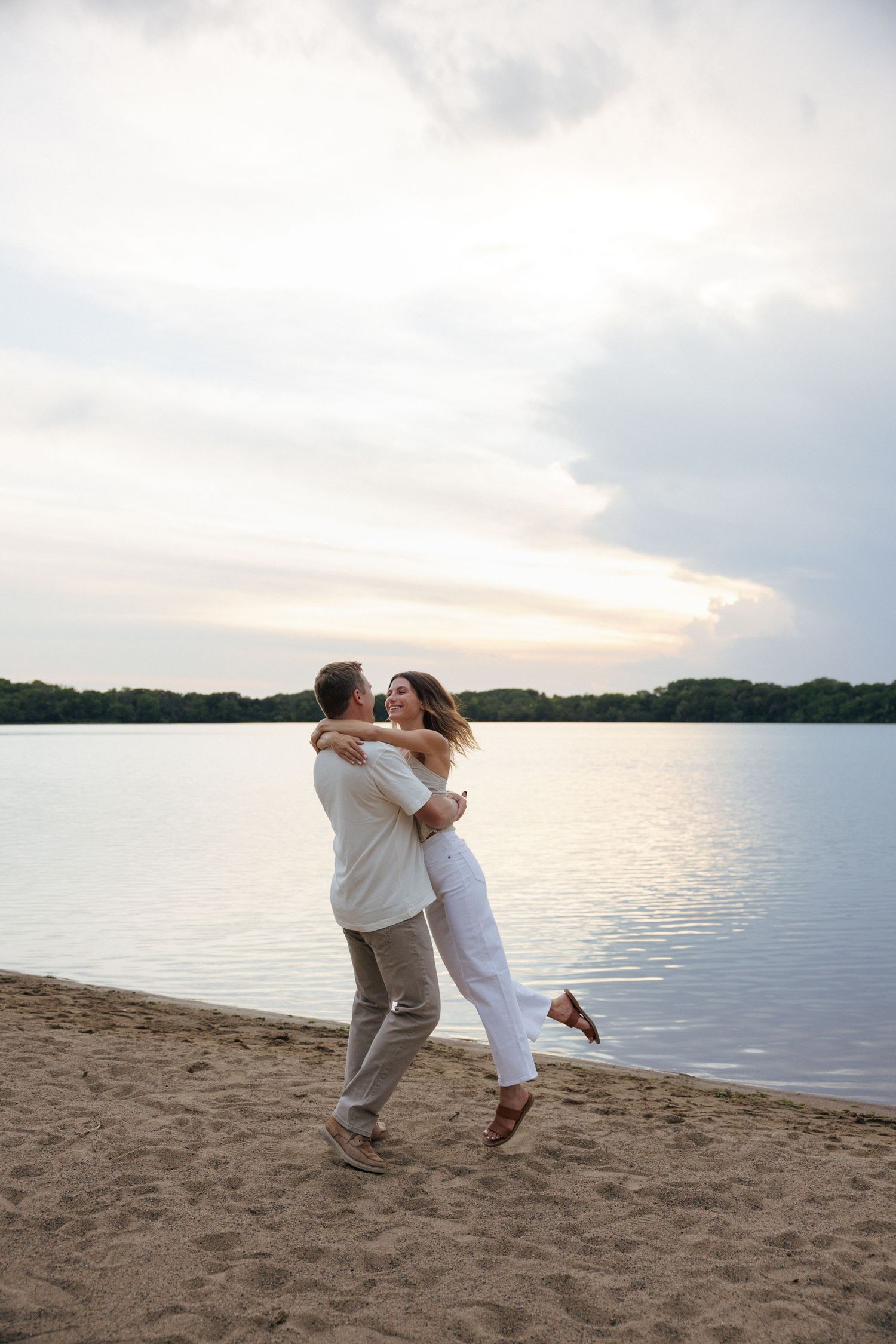 Couple embracing and dancing on a sandy beach at sunset with calm water and trees in the background.
