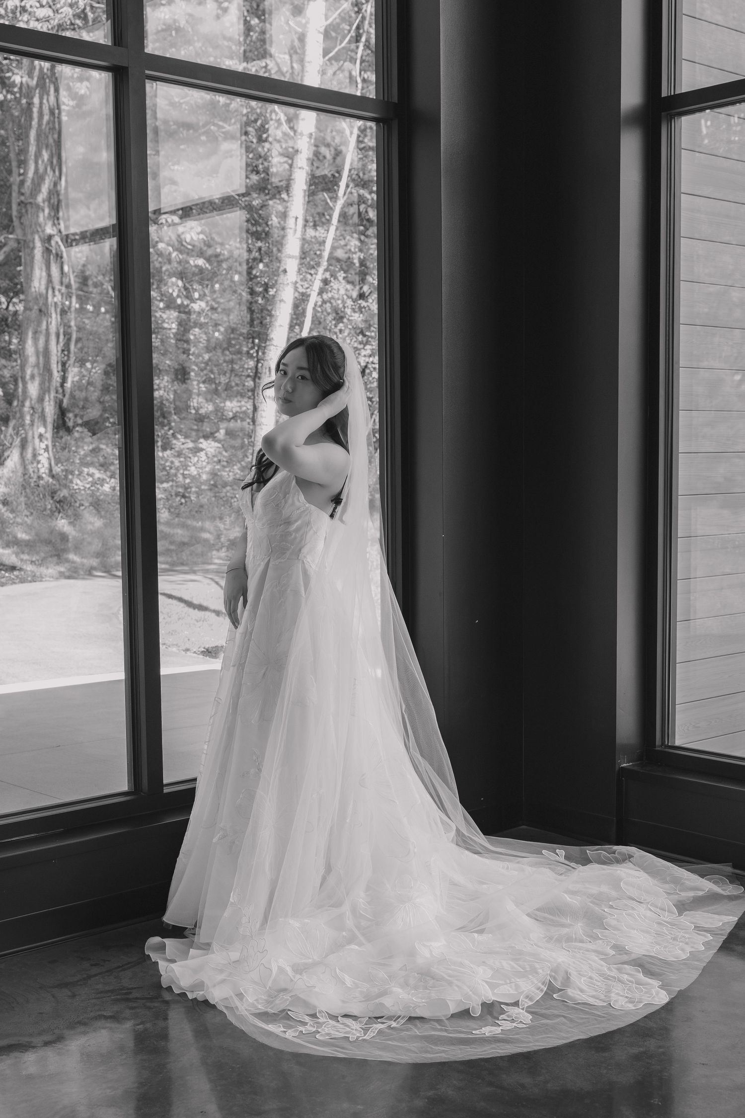 Black and white image of a bride in a lace gown and cathedral veil posing by tall windows.