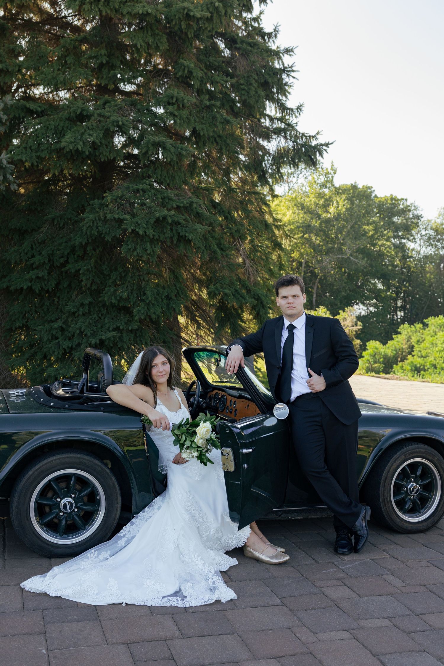 Bride in lace gown and groom in black suit posing beside a classic dark green convertible on wedding day.