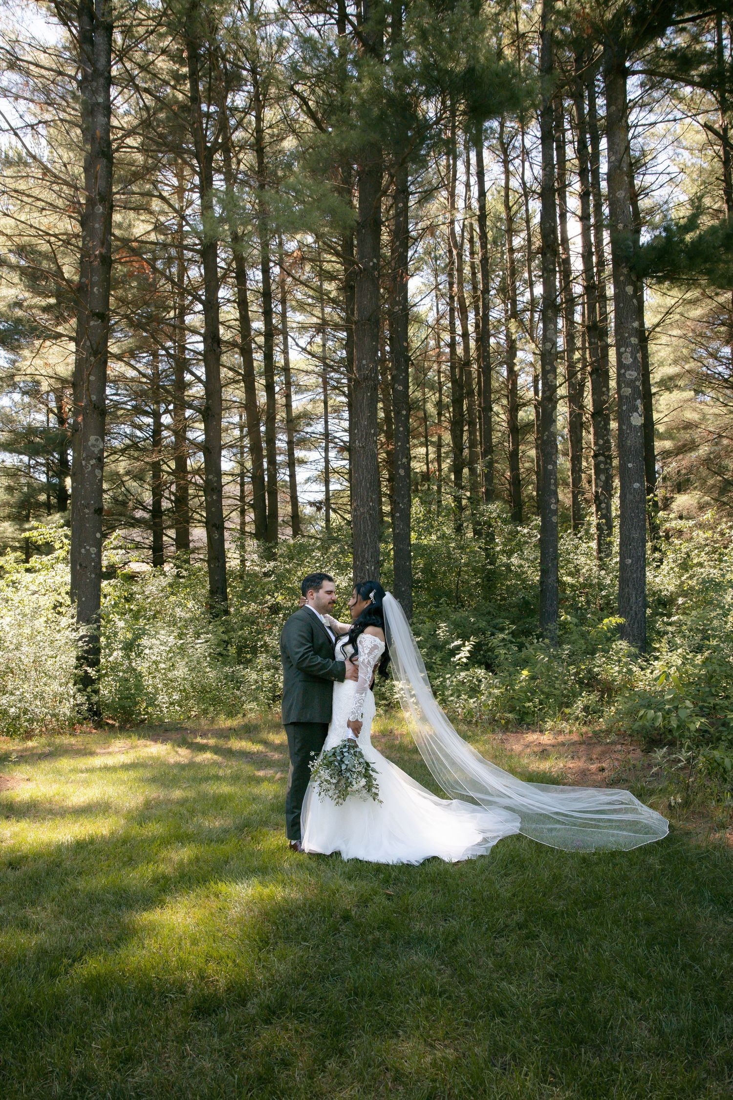 Bride and groom share a romantic moment in a lush pine forest, her veil flowing gracefully in the breeze.