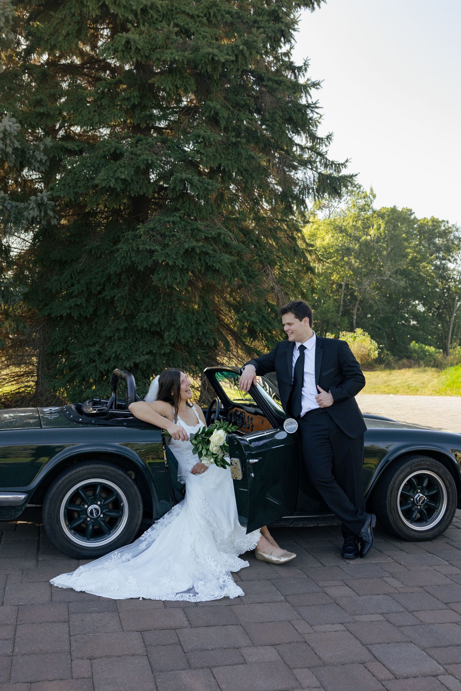Bride and groom posing beside a vintage dark green convertible car outdoors, surrounded by tall evergreen trees.