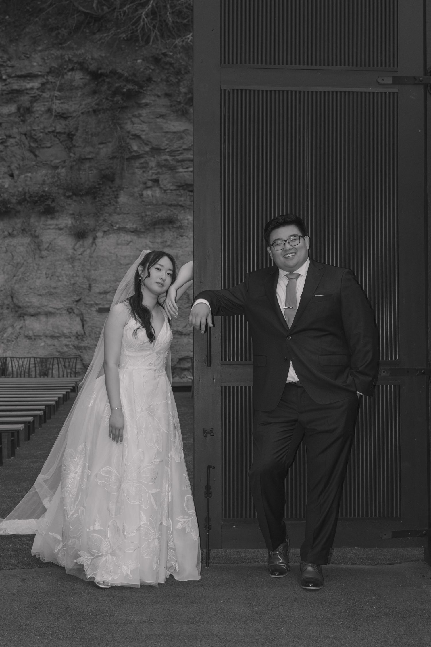 Black and white photo of bride and groom posing casually outside a chapel with stone walls and wooden doors.