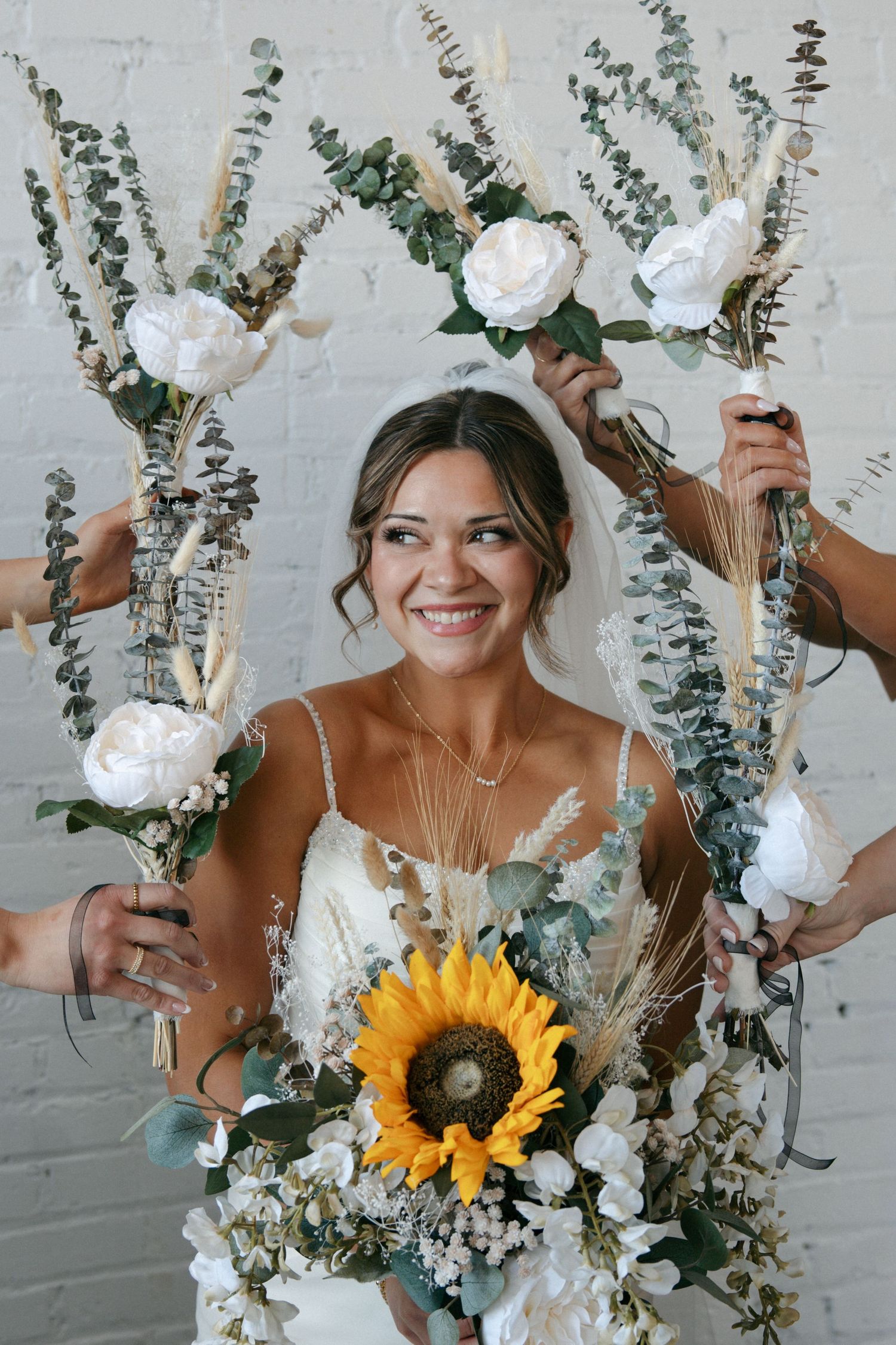 Smiling bride holding sunflower bouquet surrounded by bridesmaids raising white floral arrangements against white brick wall.
