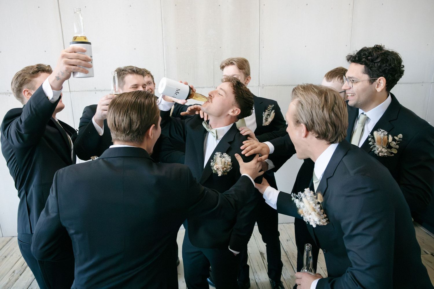 Groomsmen in black suits celebrating with the groom drinking champagne from a bottle at a wedding reception.