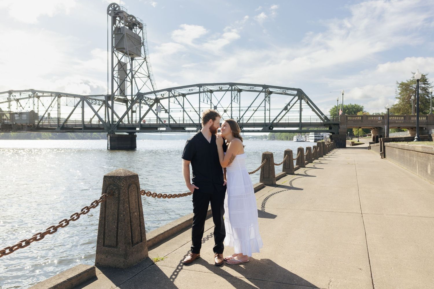 Couple embracing on waterfront promenade with historic steel lift bridge and river in background on sunny day.
