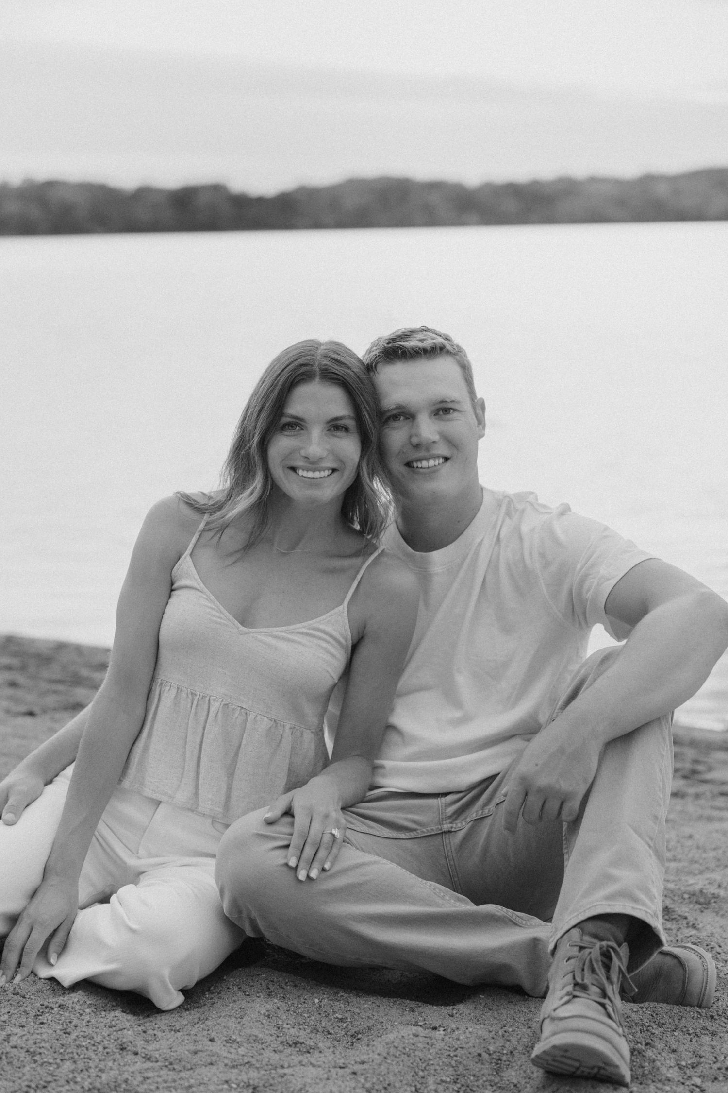 Black and white photo of a smiling couple sitting together on a sandy beach with a lake and trees in the background.