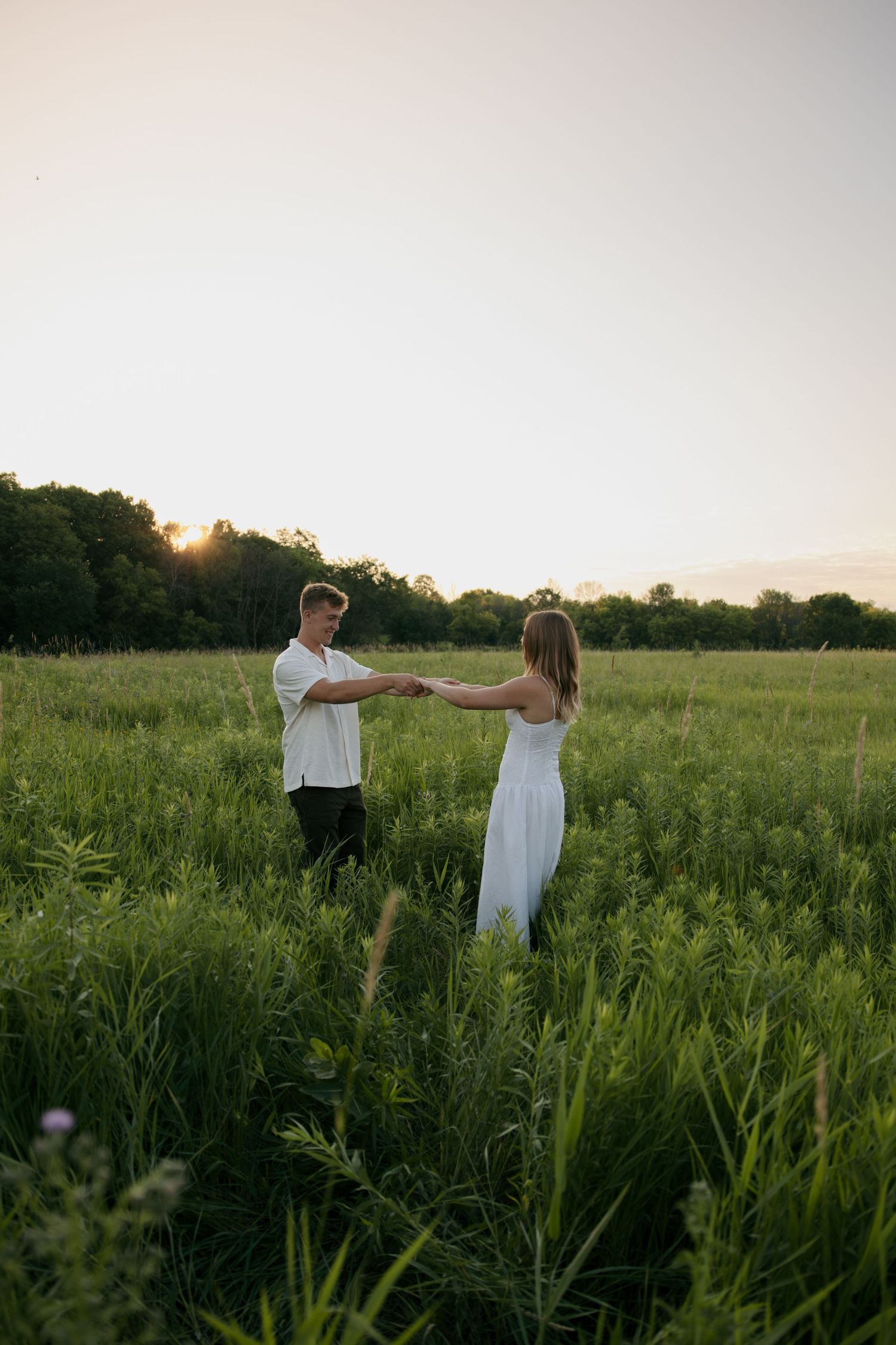 Couple holding hands in a vast green meadow at sunset, facing each other romantically.