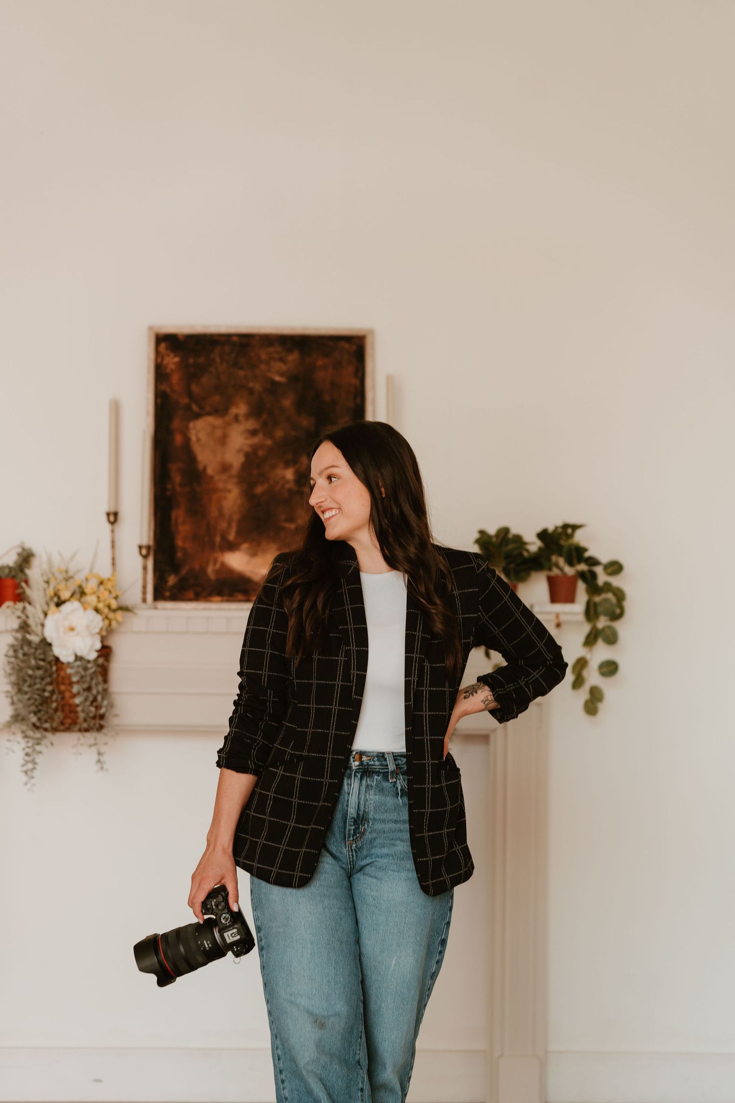 Woman photographer in plaid blazer and jeans holding camera, smiling in bright studio with art and plants.
