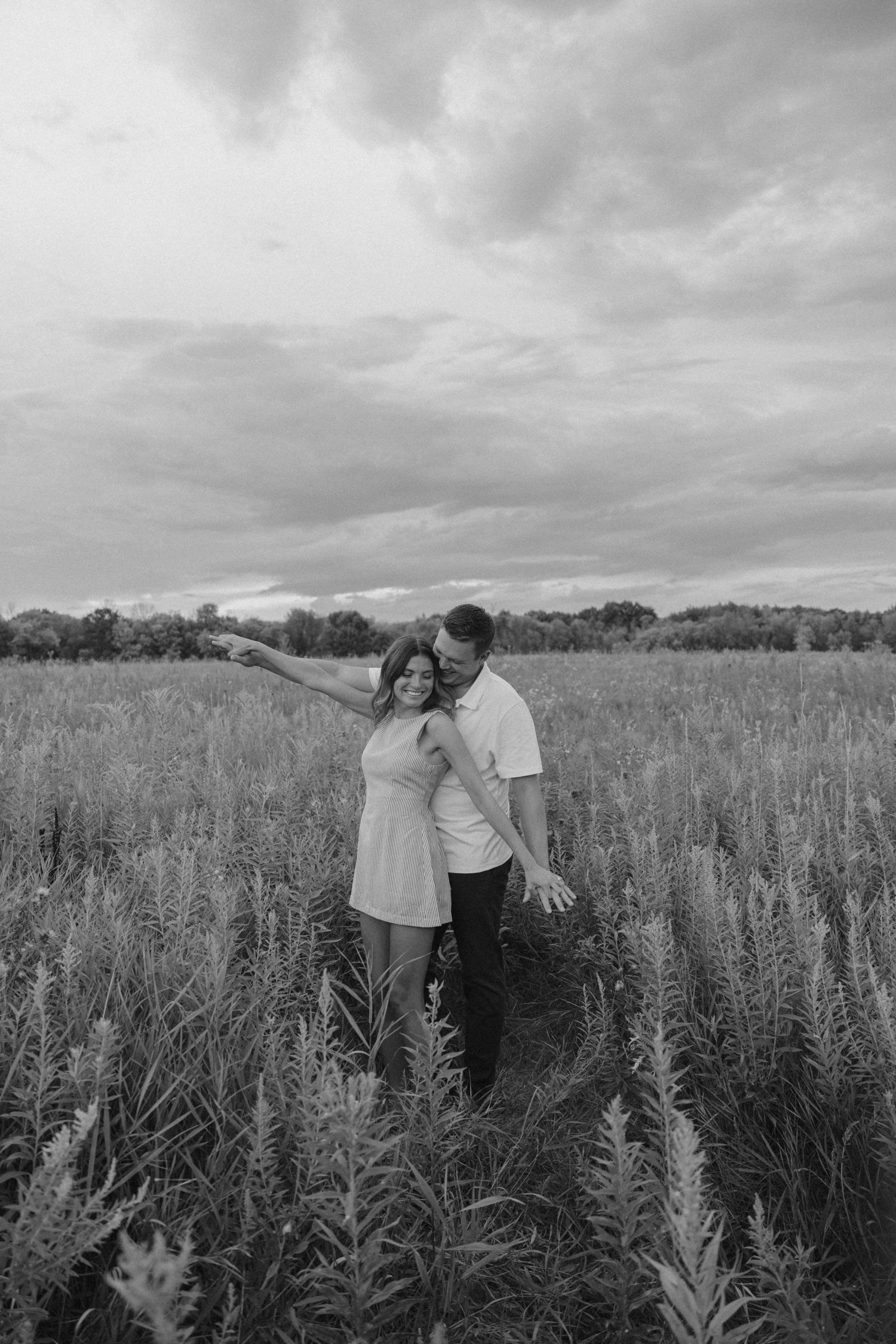 Black and white photo of a couple laughing together in a wildflower field, woman pointing.