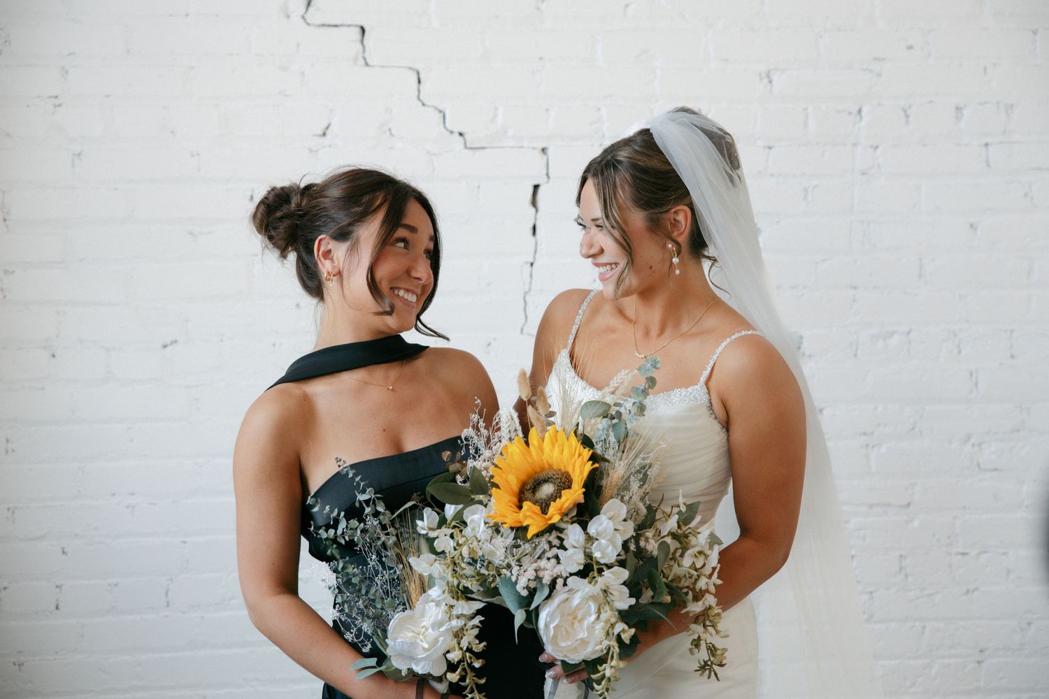 Bride in white gown holding sunflower bouquet smiles with bridesmaid in black dress against white brick wall.